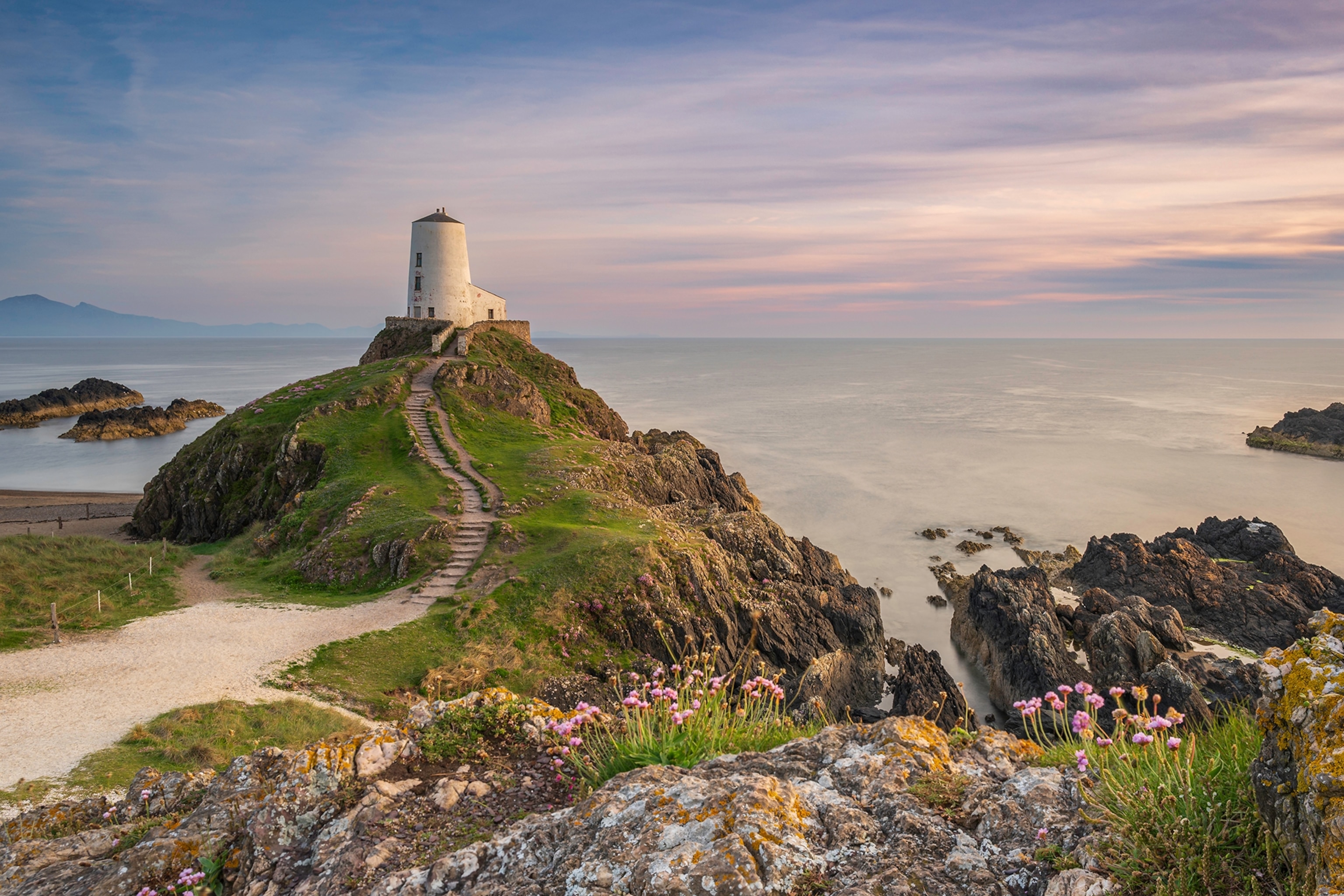 A white lighthouse on a rocky spit with steps leading down through the grass and the sea behind it.