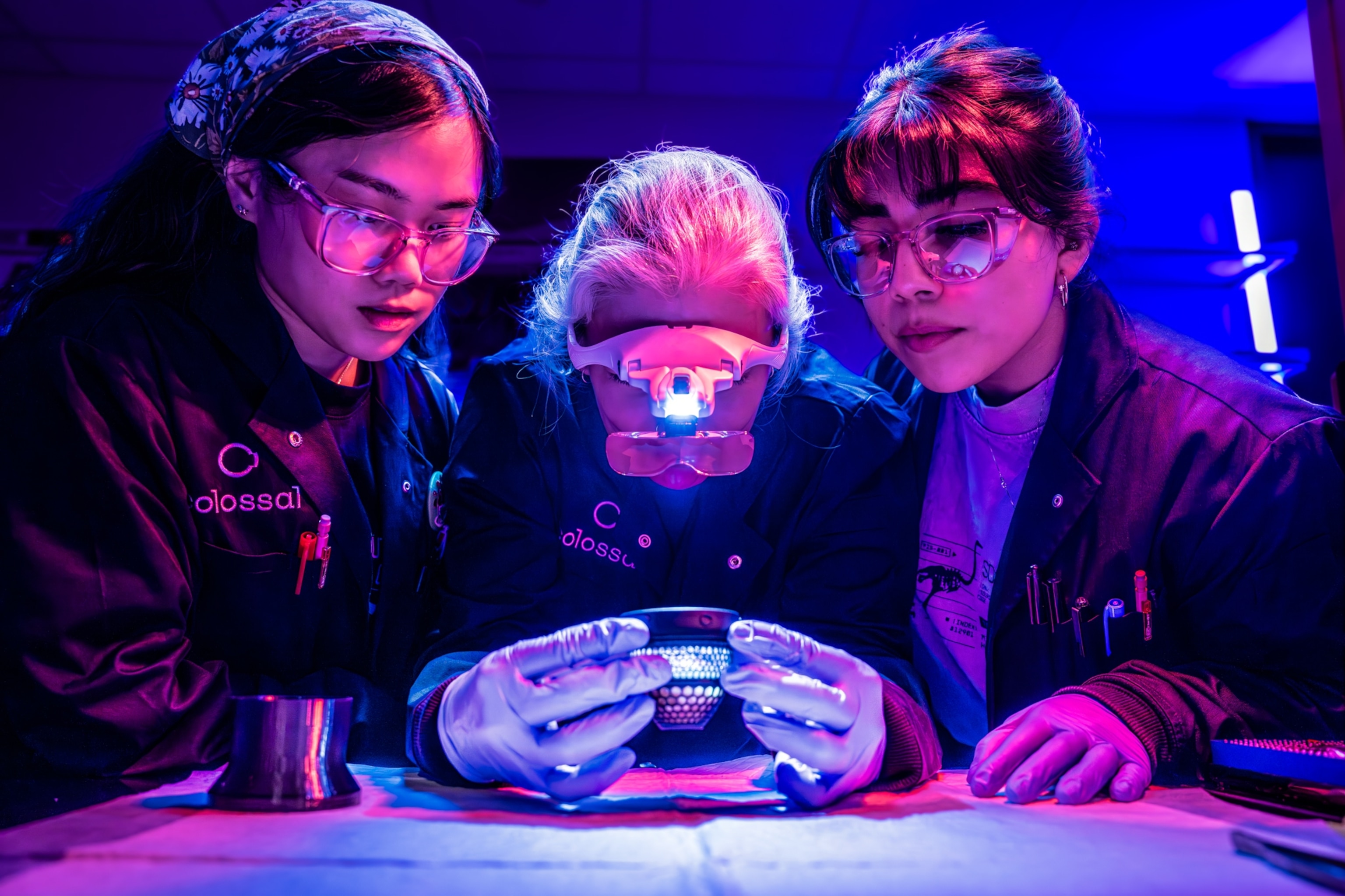 Three scientists in a lab examine part of an artificial egg under blue and purple lighting