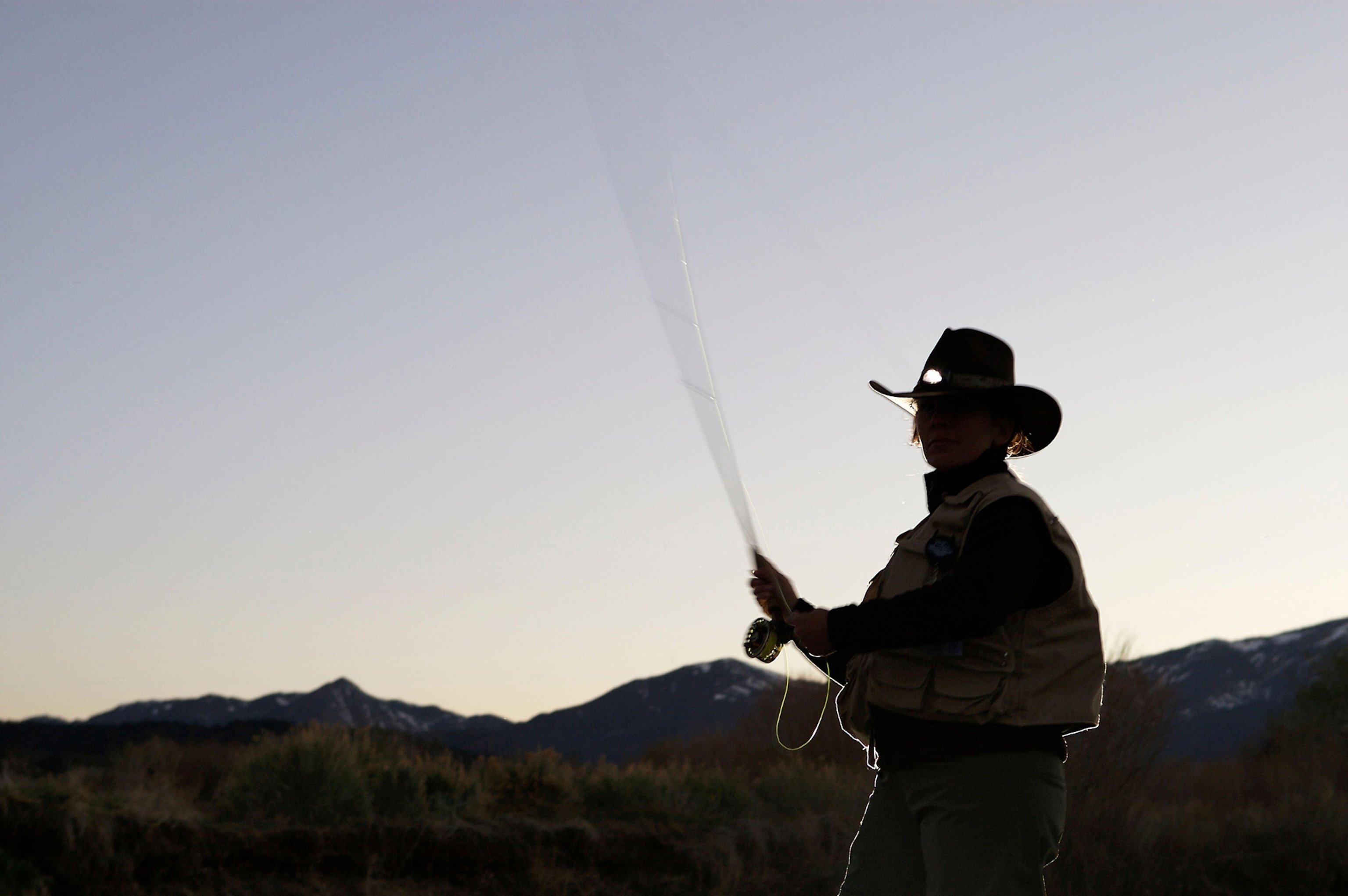 A woman holding a fly fishing rod and wearing a cowboy hat is silhouetted against a dusky blue sky