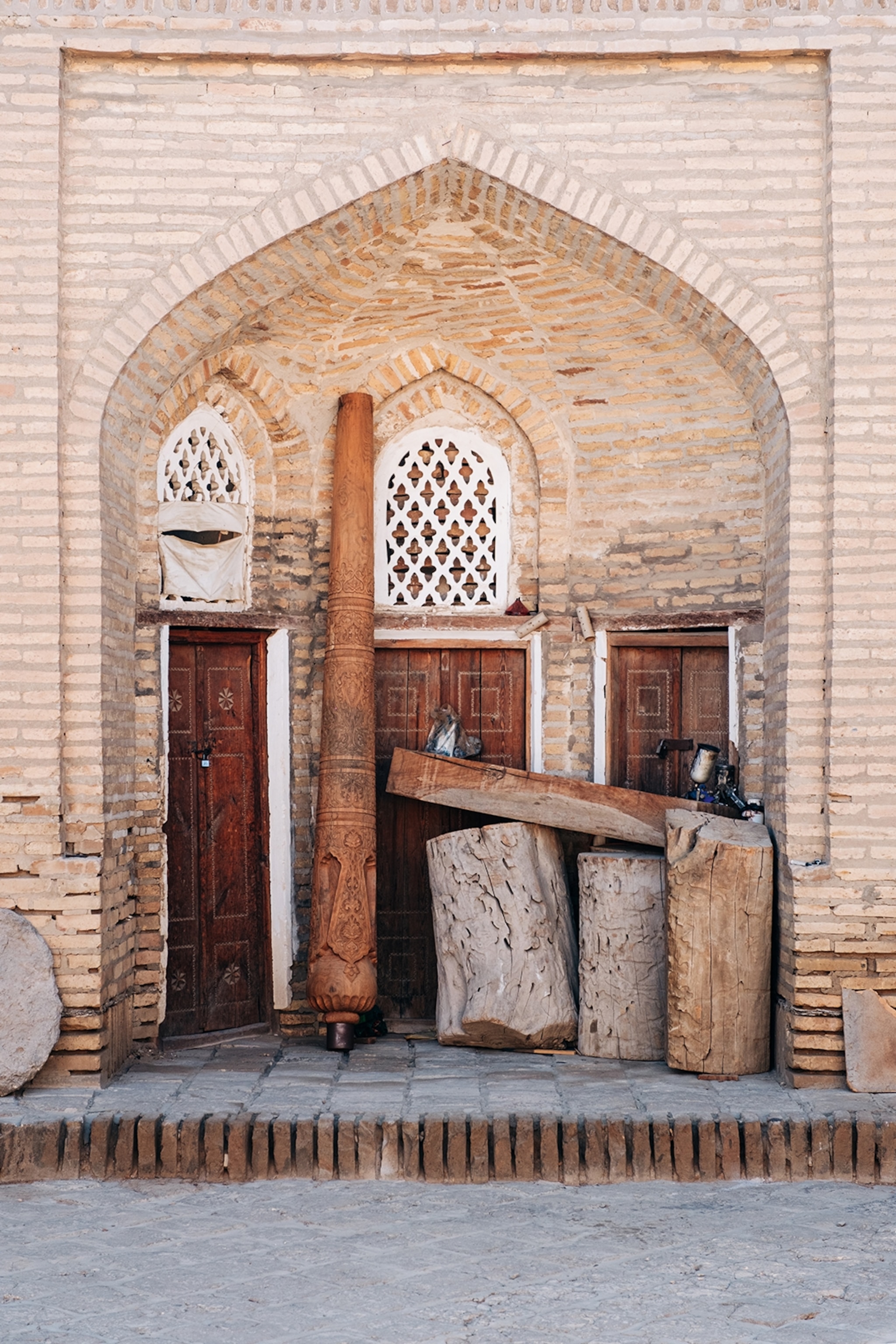 The entrance to a stone building with wooden logs and columns leaning against two of the three tall wooden doors.