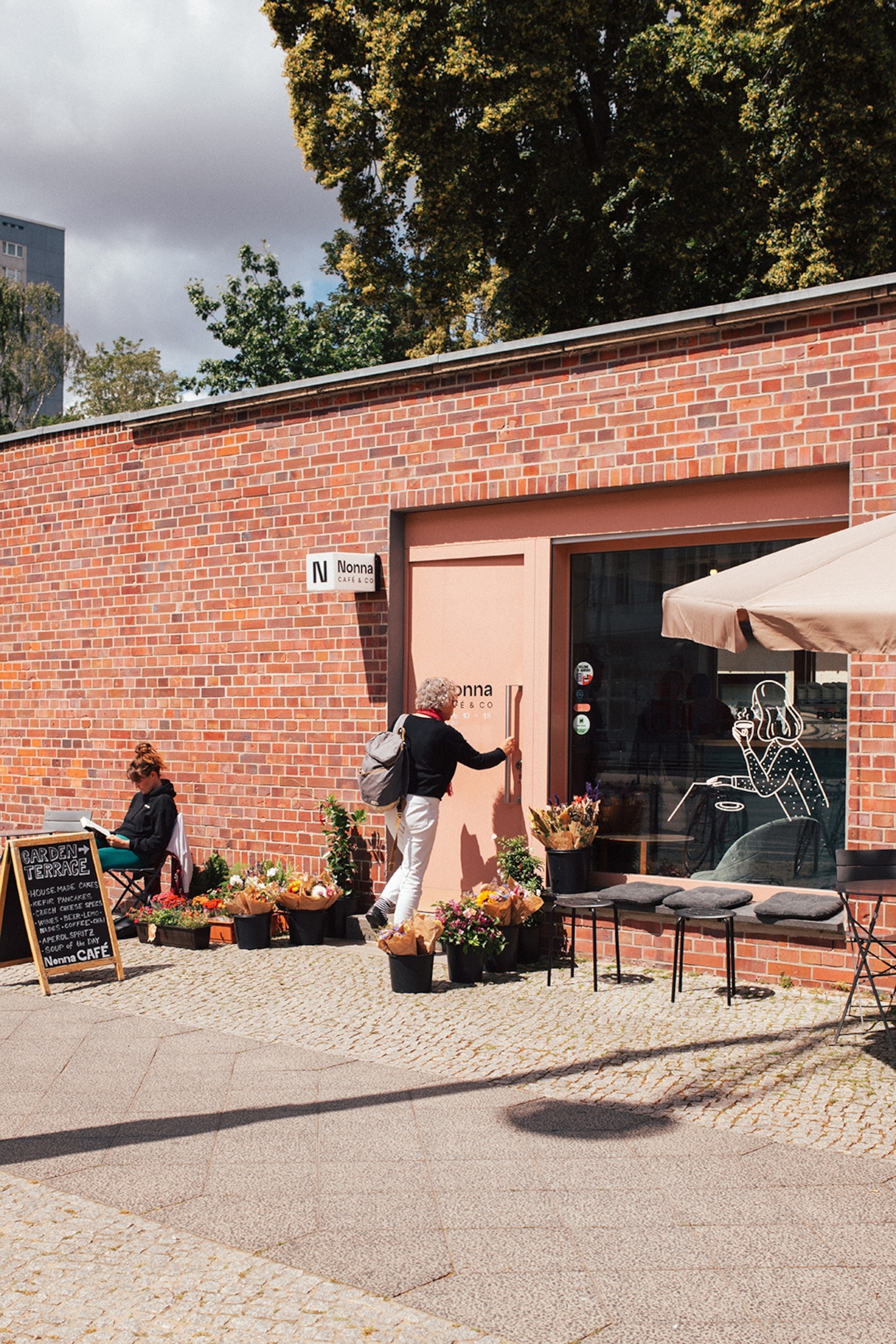 A small cafe in a red brick building with a menu board standing outside and an elderly woman entering through the glass entrance.