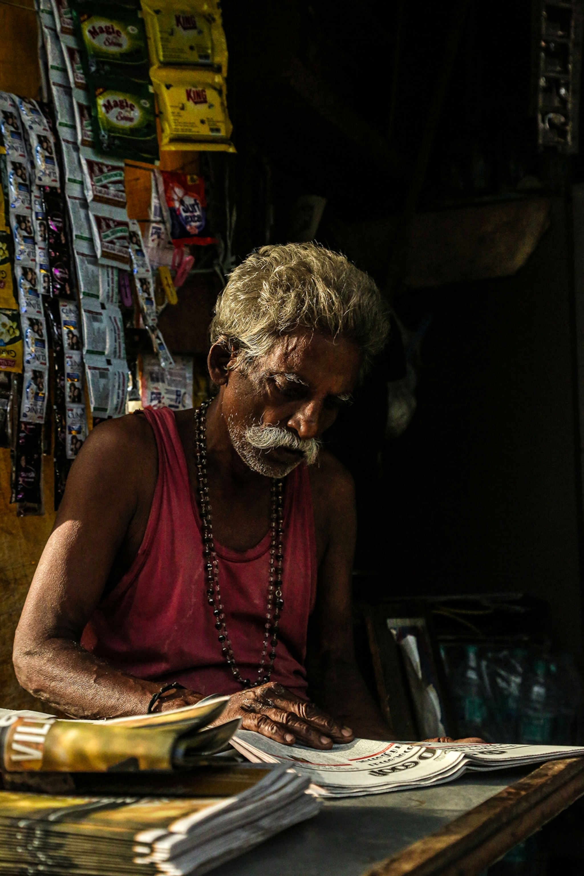 An elderly man sitting inside a shop reading the newspaper with light trickling in through a window.