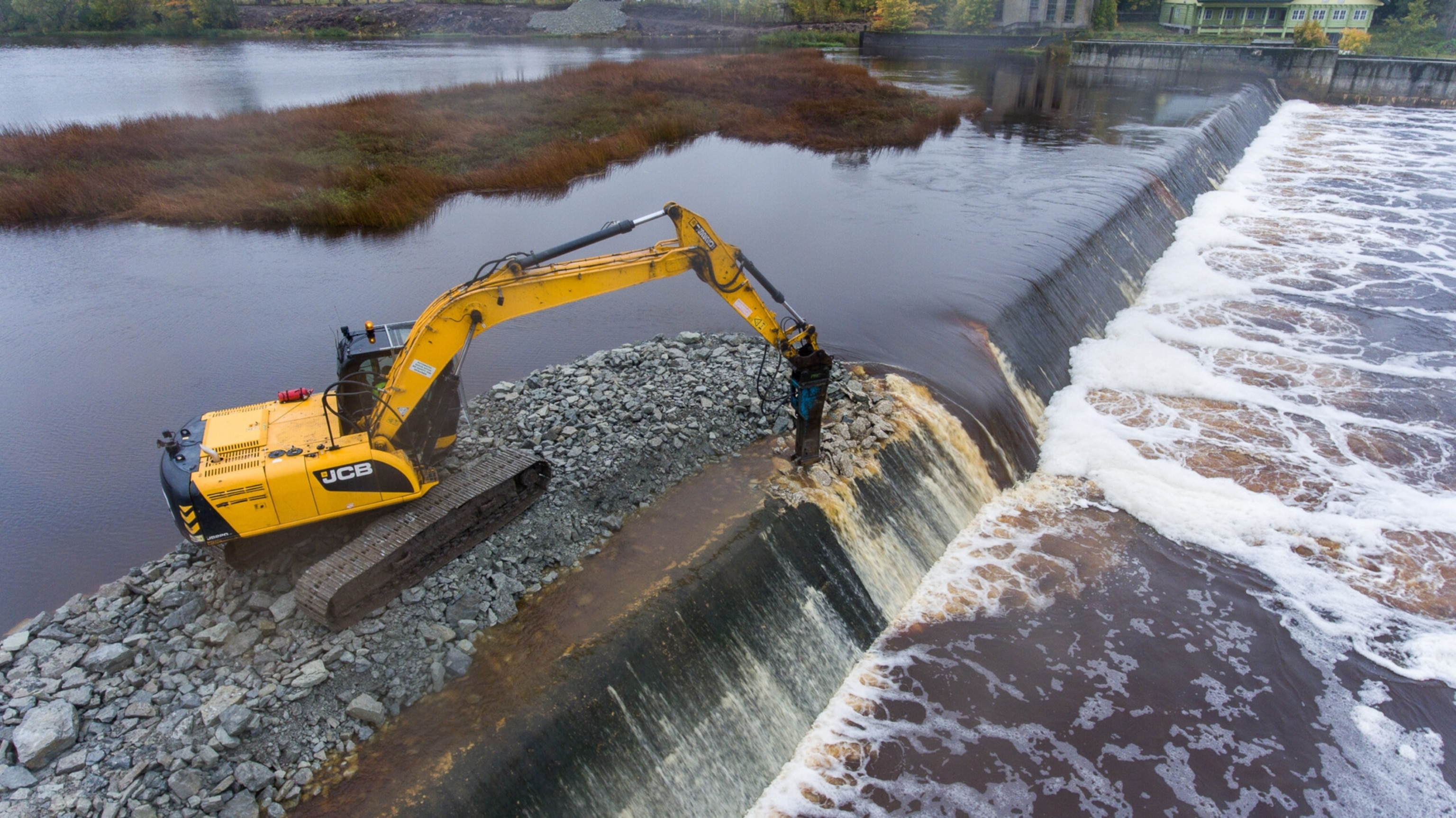 A yellow excavator on a rock path with its arm extended towards water flowing over a short dam