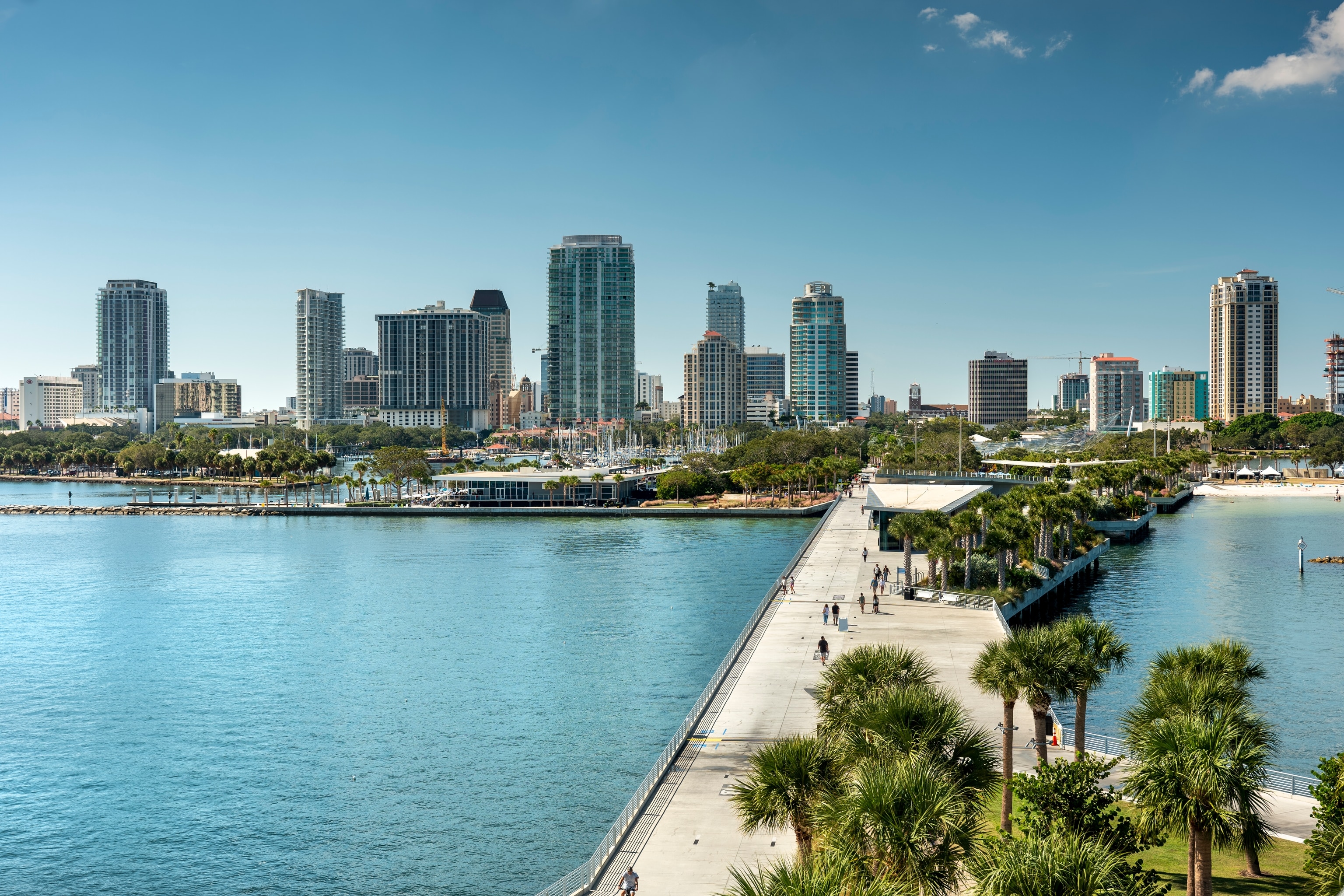 Concrete boardwalk lined with palm trees extends from the sea towards a city skyline of high-rise buildings, boats moored in a harbour and a white-sand beach dotted with umbrellas