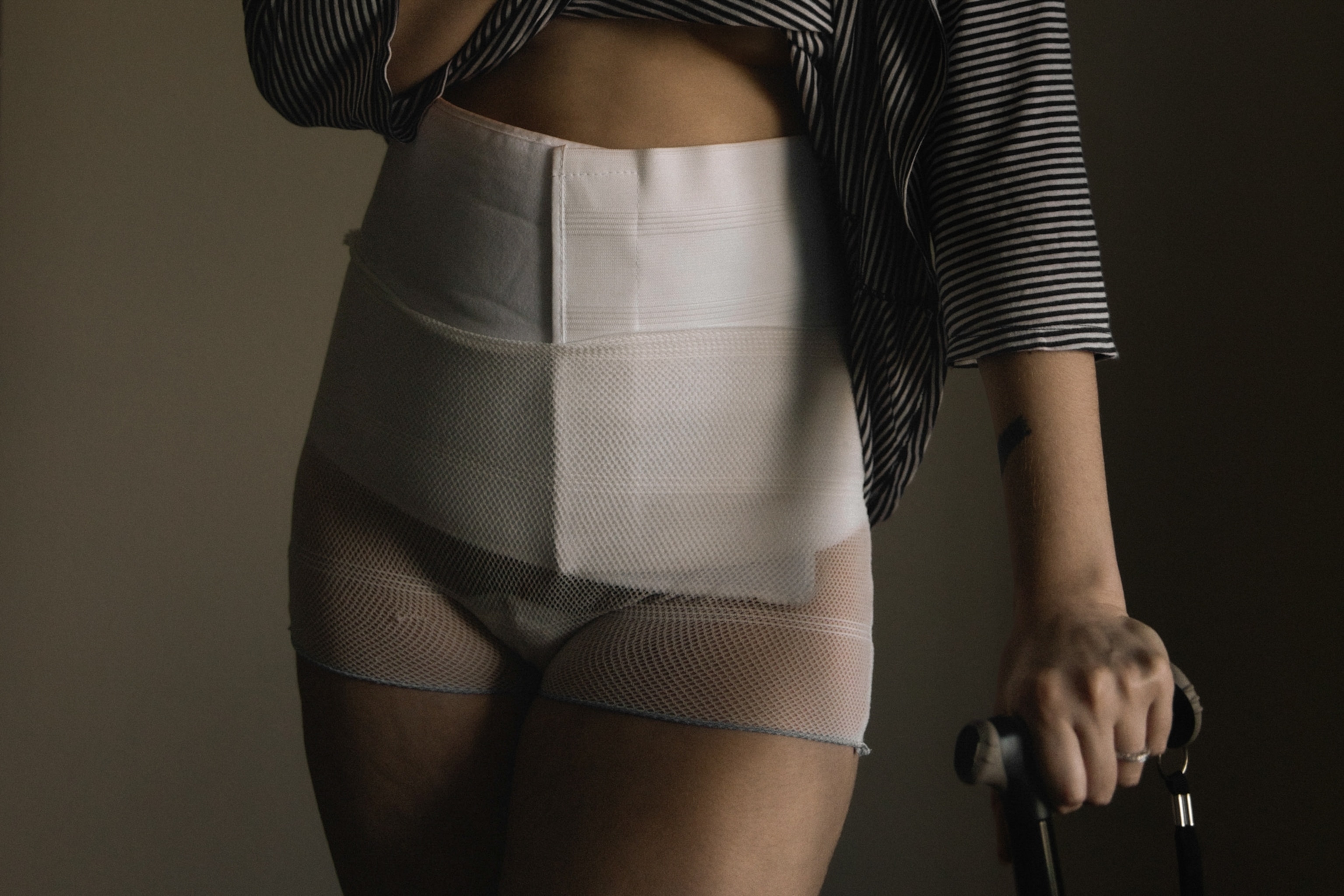 Close-up of a person wearing a striped shirt, white abdominal binder, white medical compression shorts, and holding a black cane.