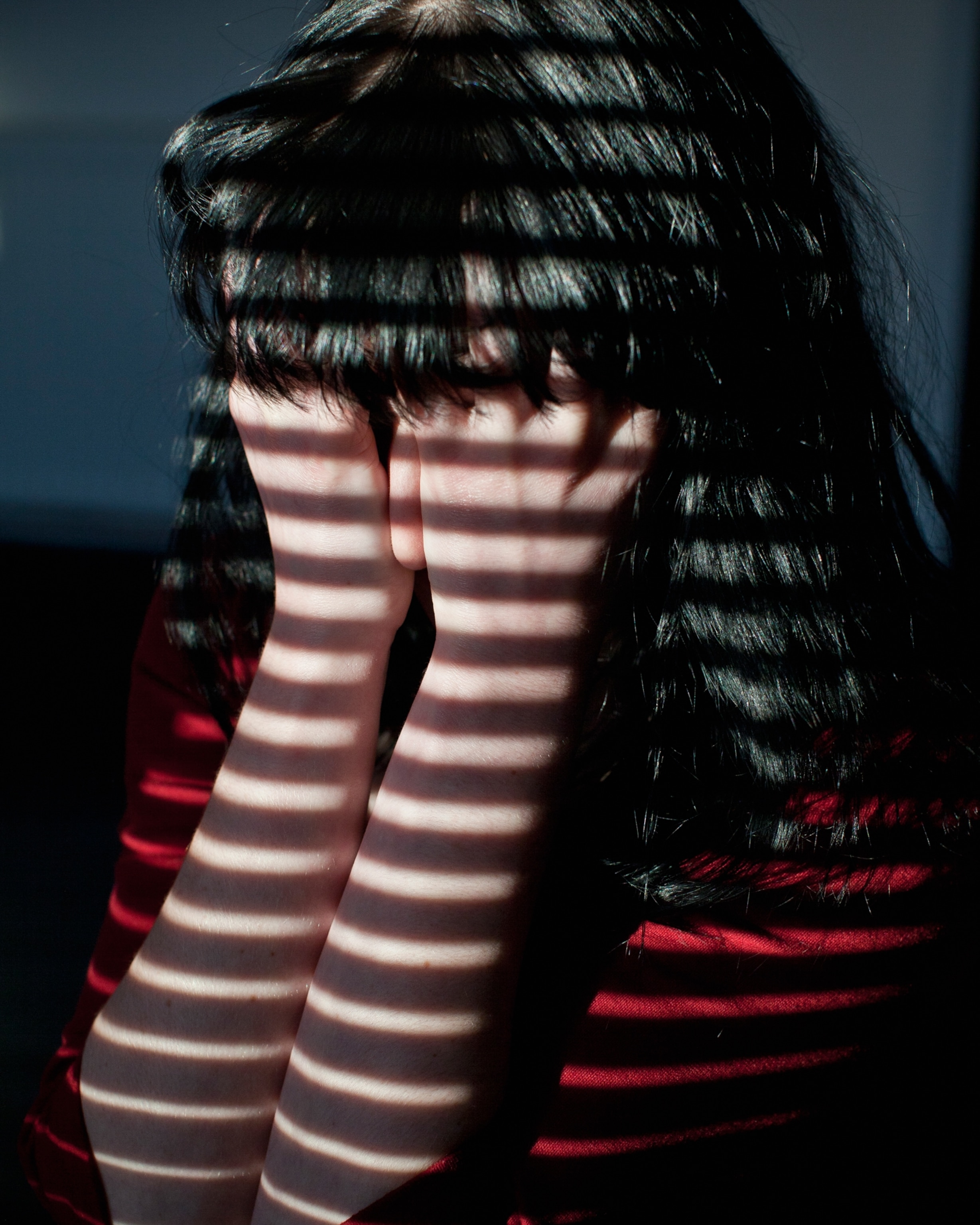 A person covers their face with their hands, illuminated by striped shadows from blinds.