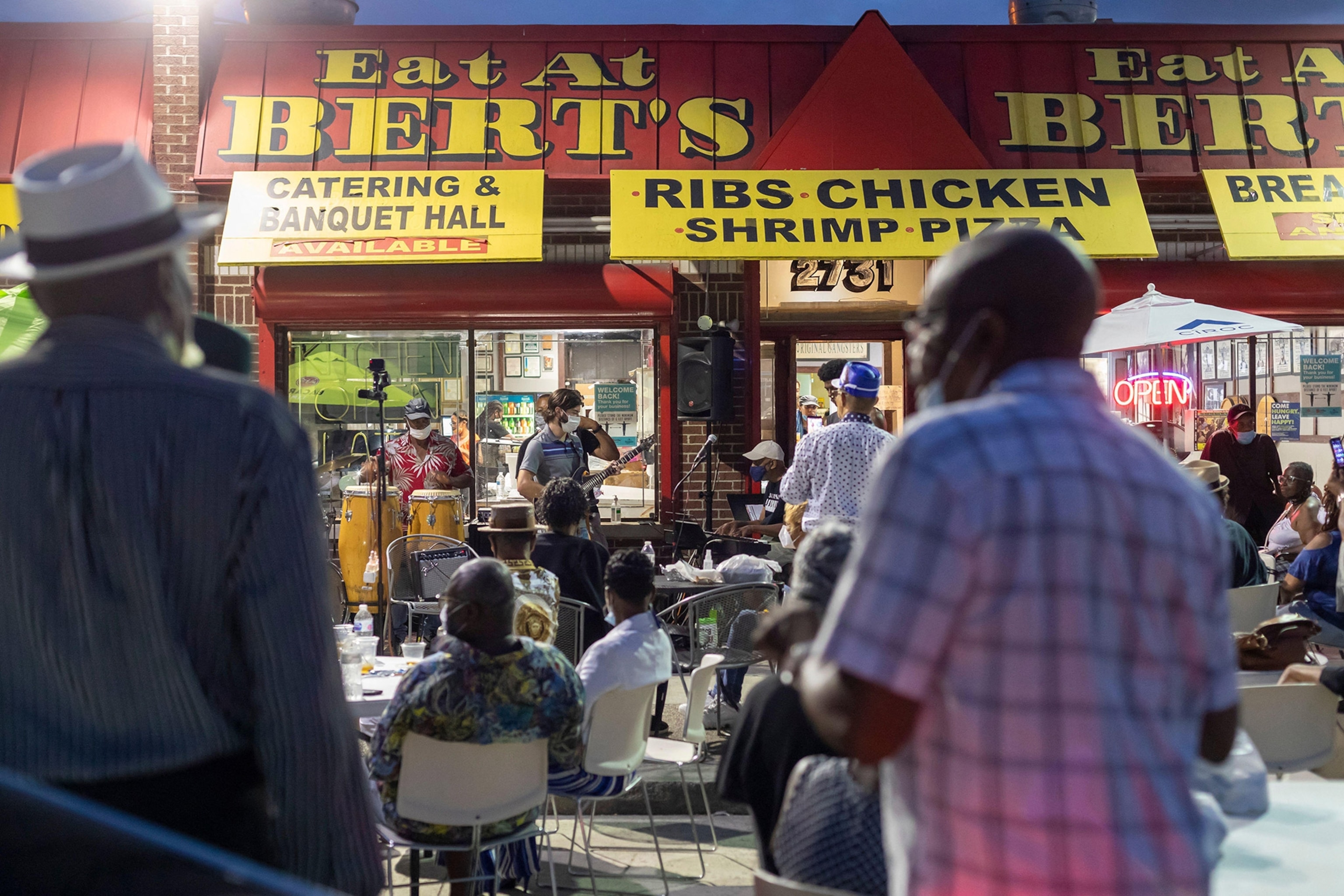 A crowd gathers in front of building with yellow signage reading EAT AT BERT'S, as musicians perform