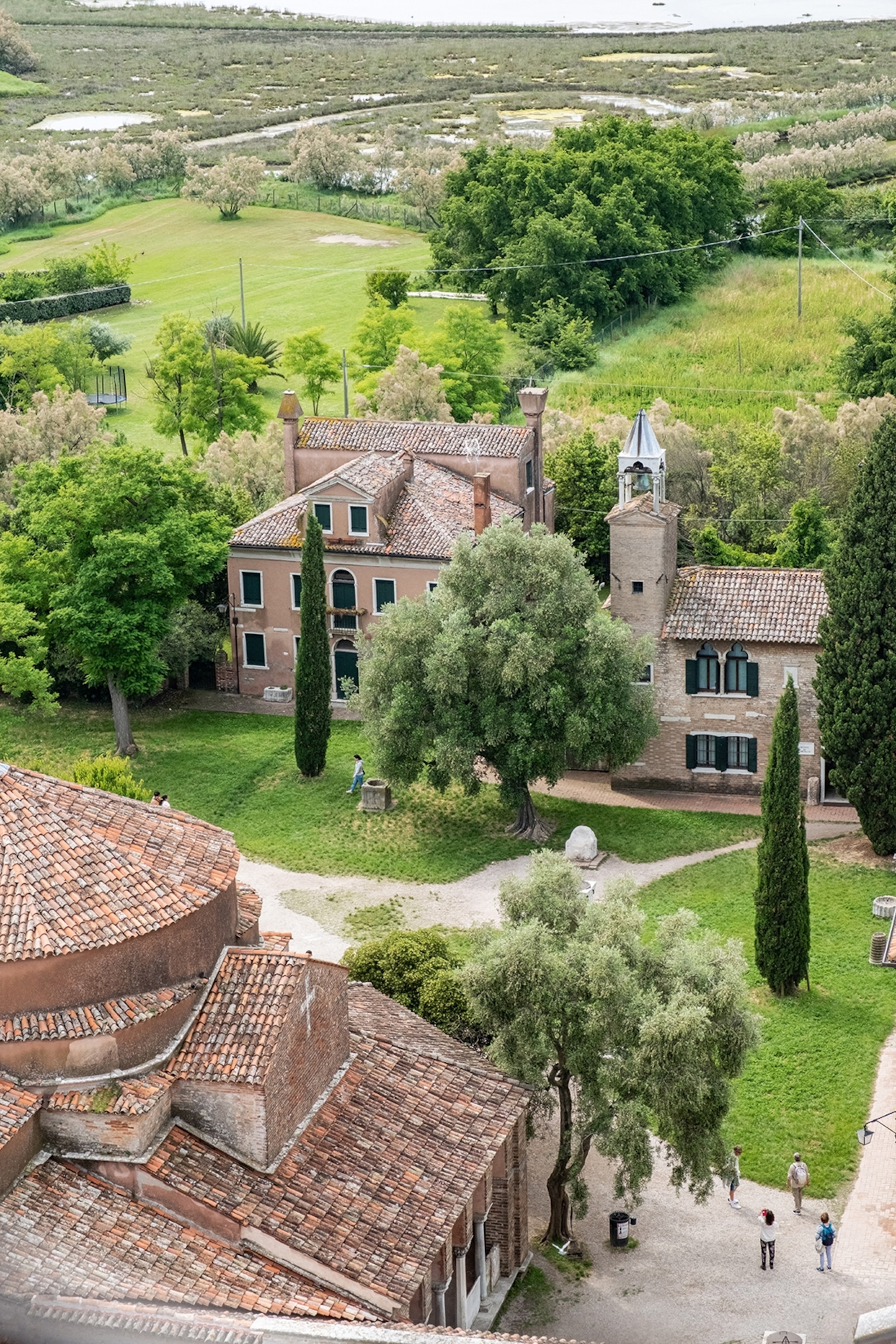 The view from a tall tower onto the grounds of an Italian estate of stone houses, with cypress tress and marshlands in the background.
