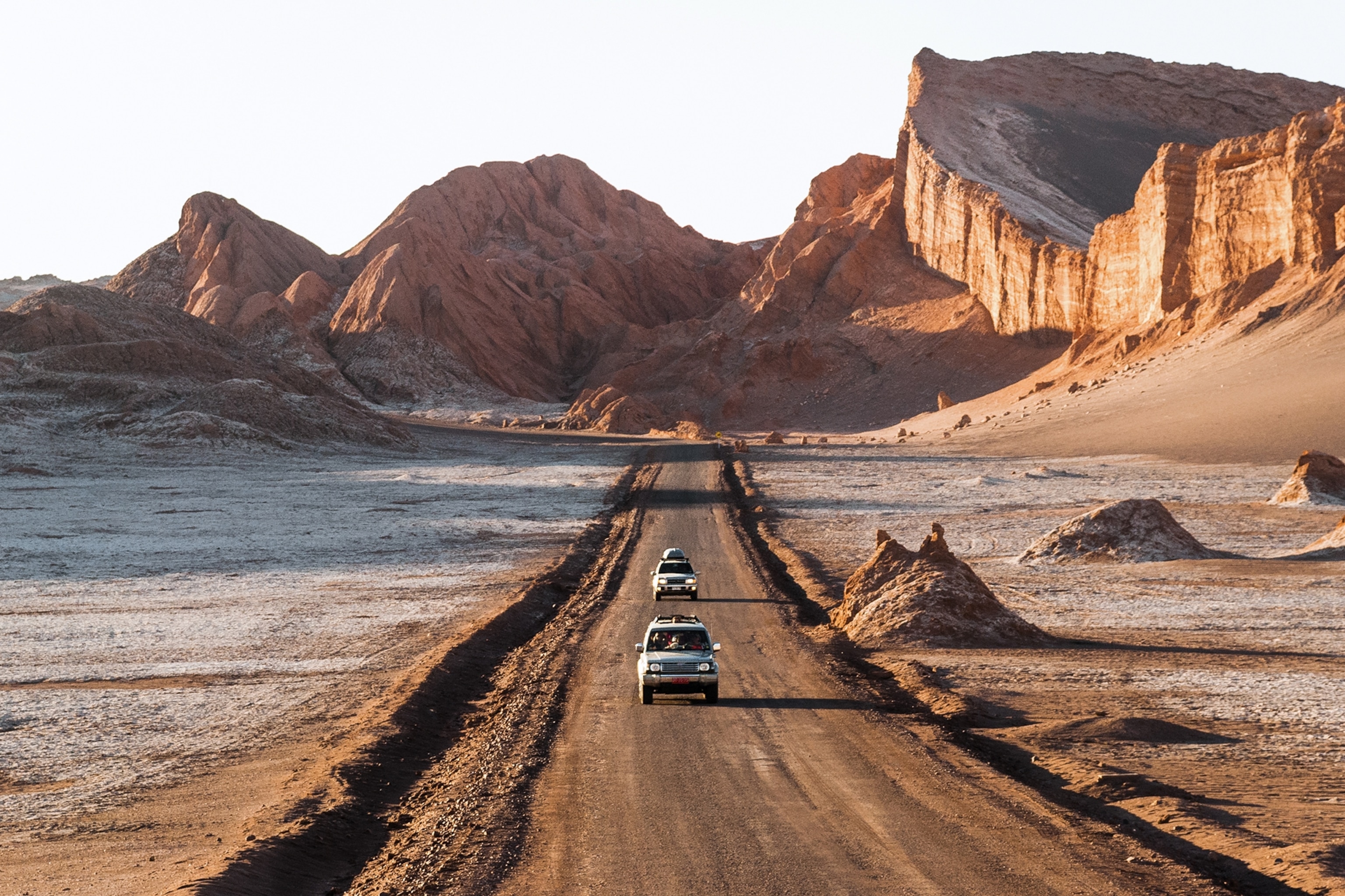 Two vans driving down a dusty desert road with impressive rock formations in the background.
