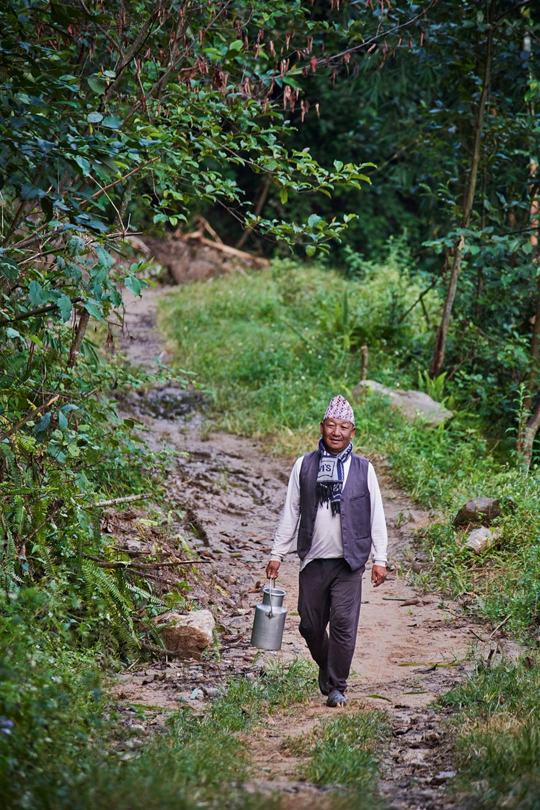 A friendly man walking down a slight hill from a forest entrance, carrying a milk churn.