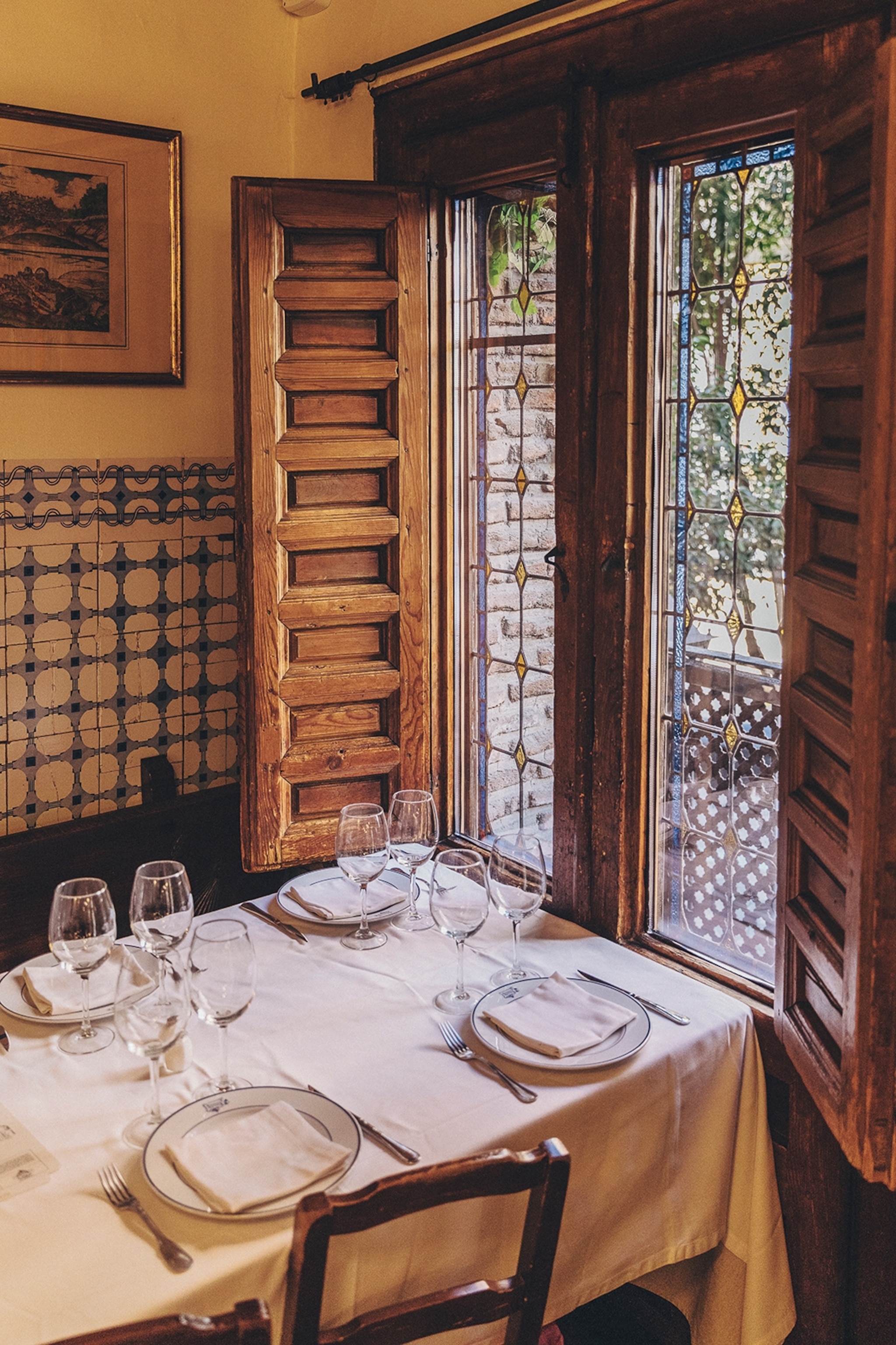 The interiors of an old restaurant with a linen-decked table pushed up to a tall window with wooden shutters opening inwards.