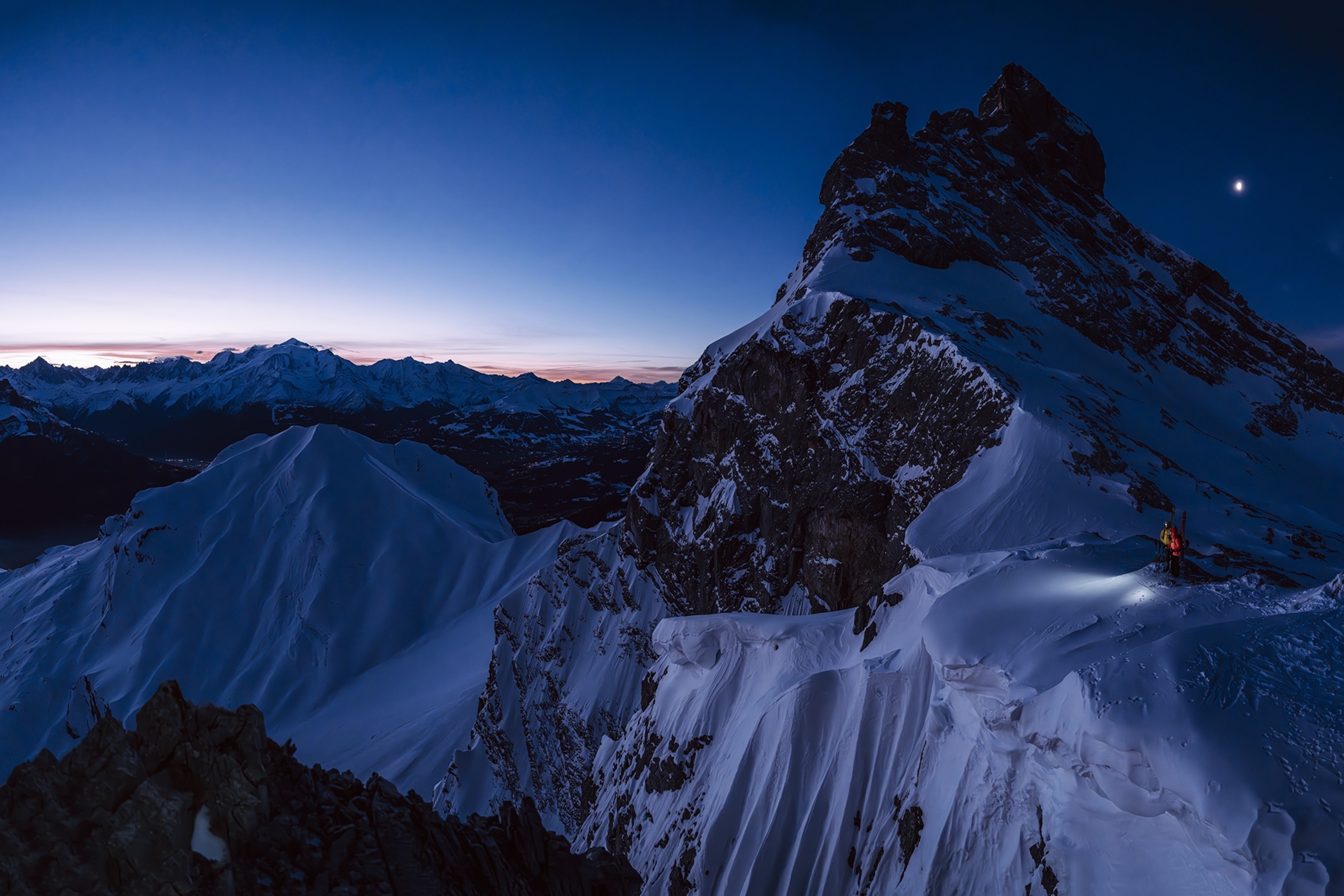 A nighttime mountain scene of a snowy peak with a few mountaineers shining their headlights into the still distance.