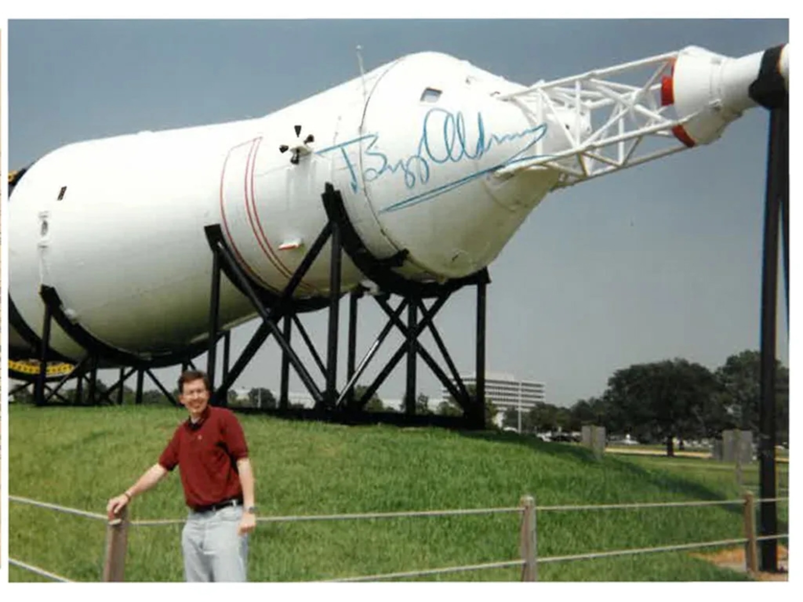 A man in a red shirt stands smiling in front of a large, white rocket displayed outdoors on black supports.