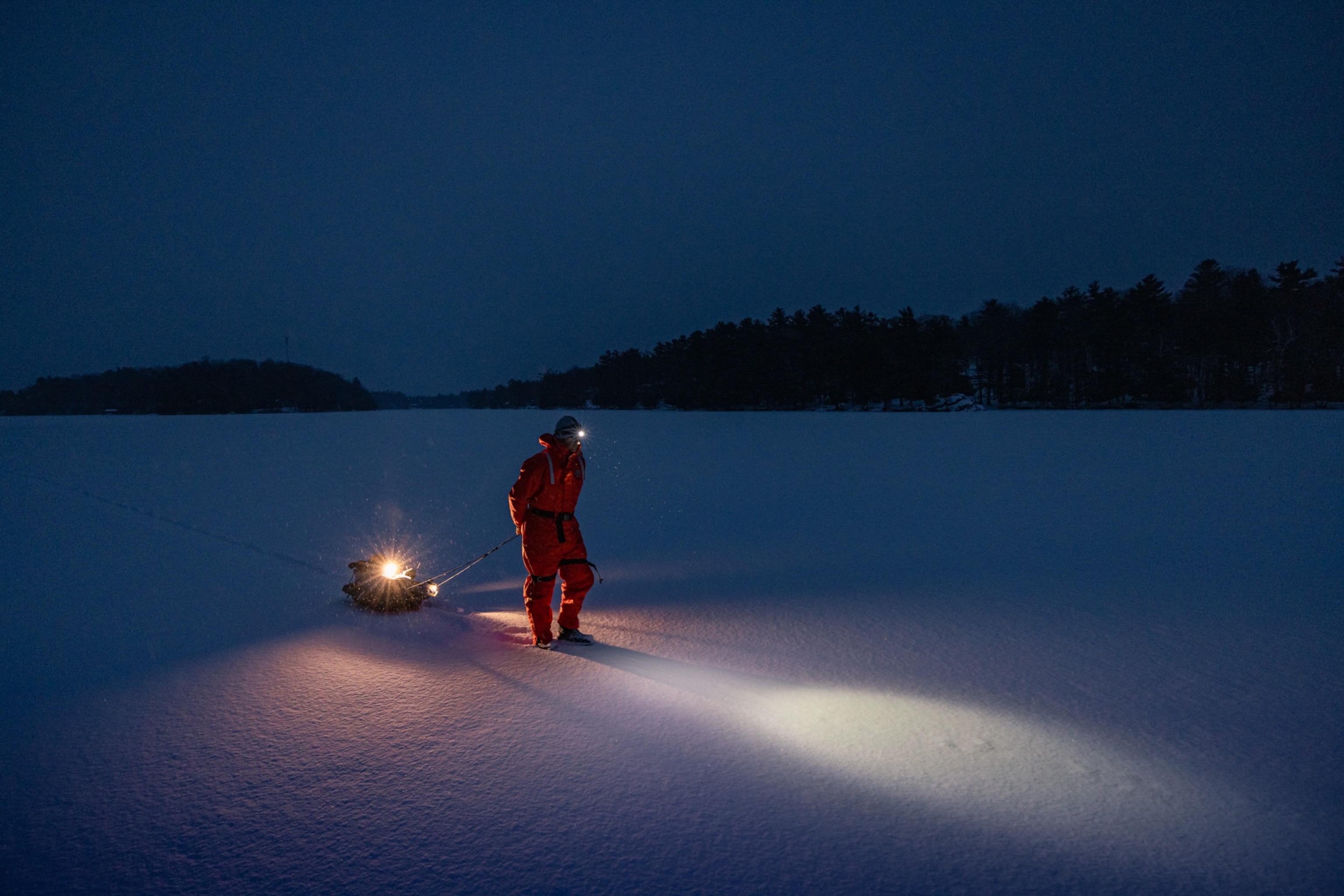 A man in an orange jumpsuit crosses a lake a night with a head lamp.