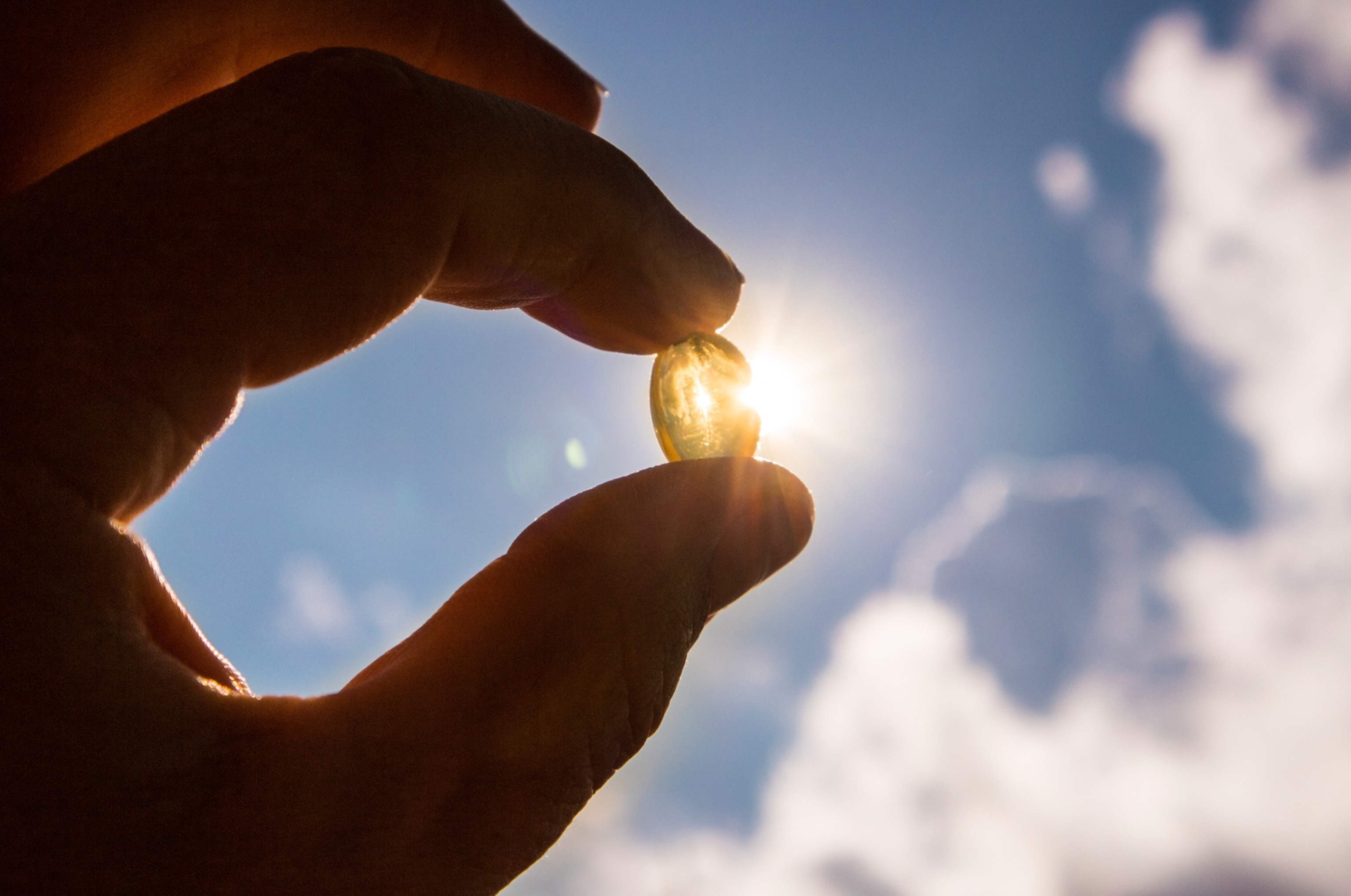 Hand holding a translucent yellow vitamin D capsule against a bright sky, sun shining through it.