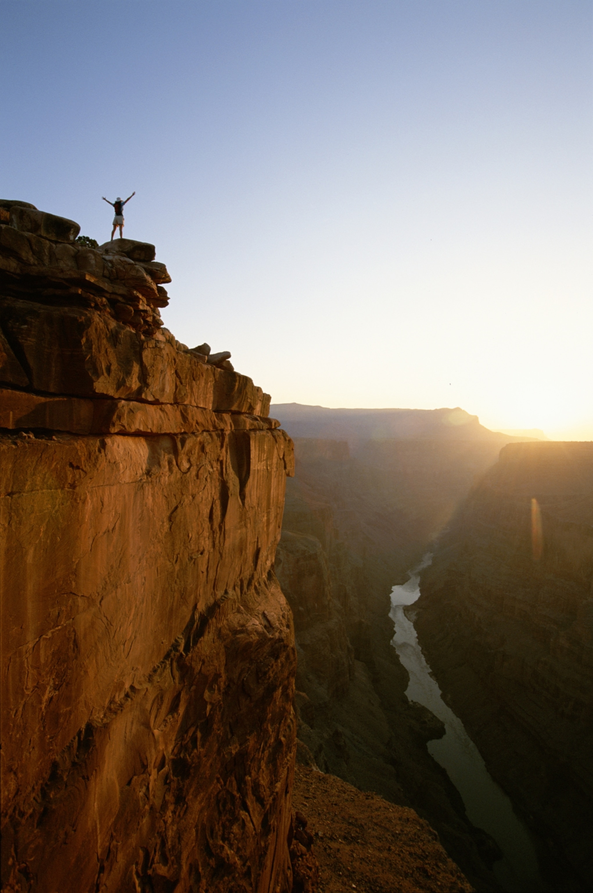 A person stands triumphantly on a cliff edge at sunrise, overlooking a vast canyon with a winding river below