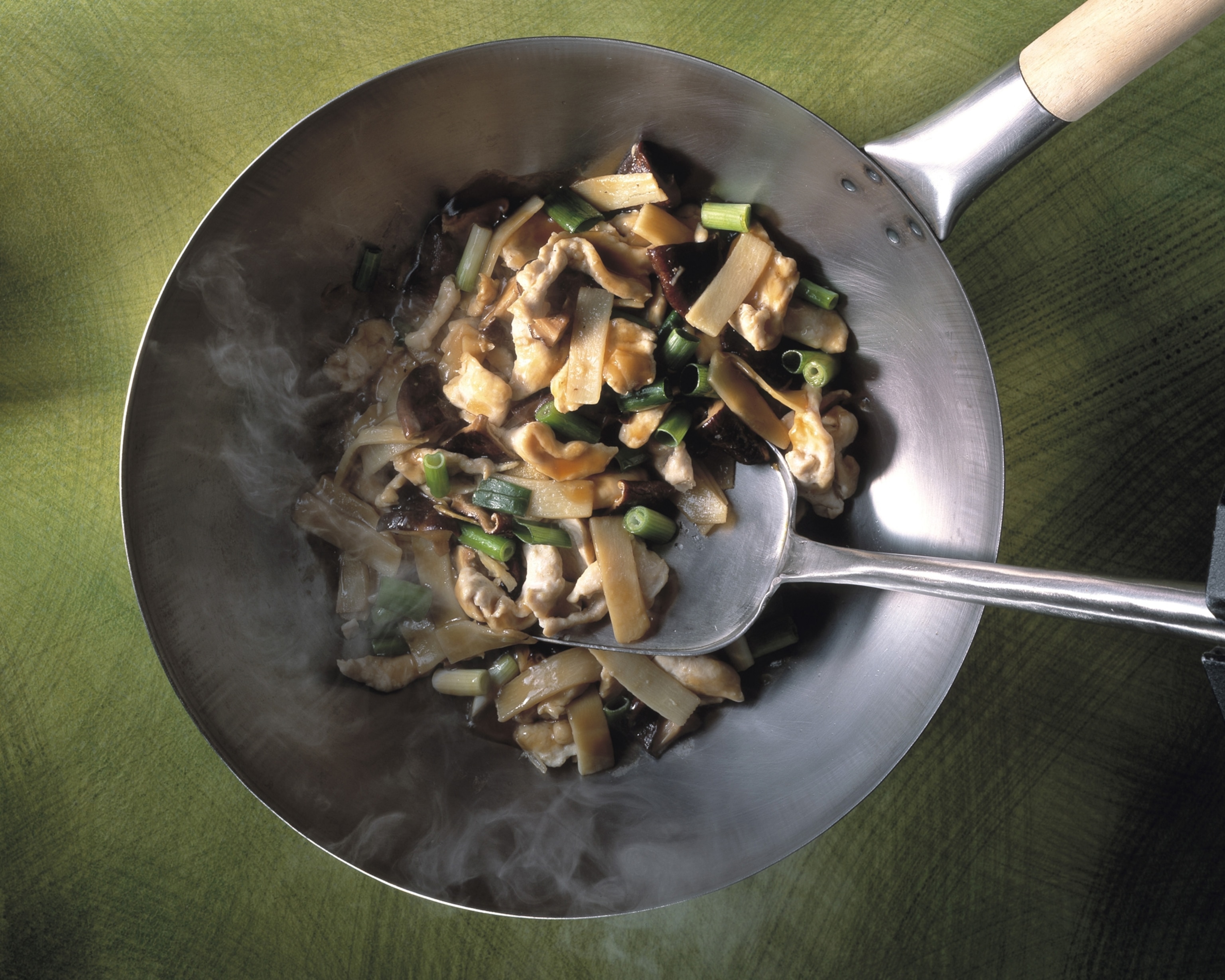 Stir-fried bamboo shoots, chicken, and green beans in a steaming metal wok with a wooden handle, set on a green surface