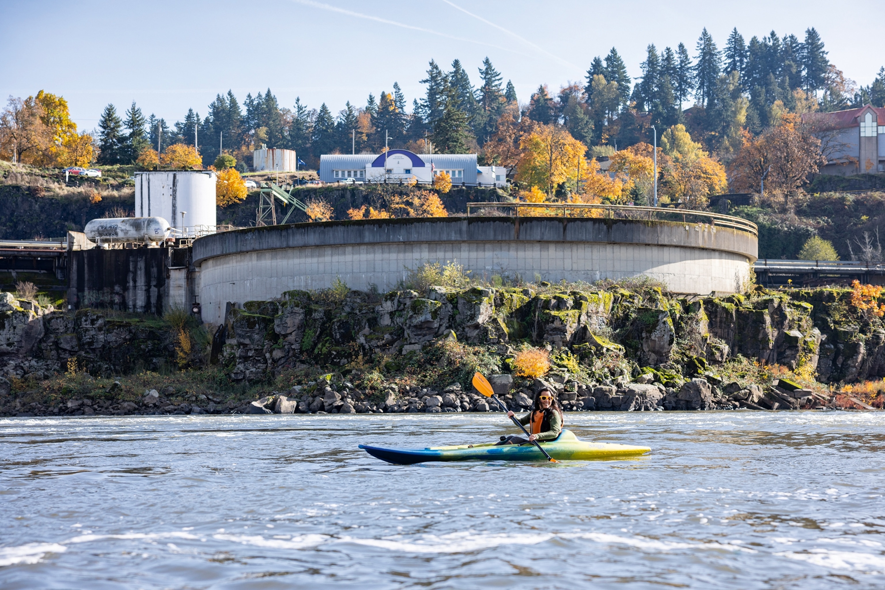 A woman smiling in a kayak and going down a river with a building and forest in the back.