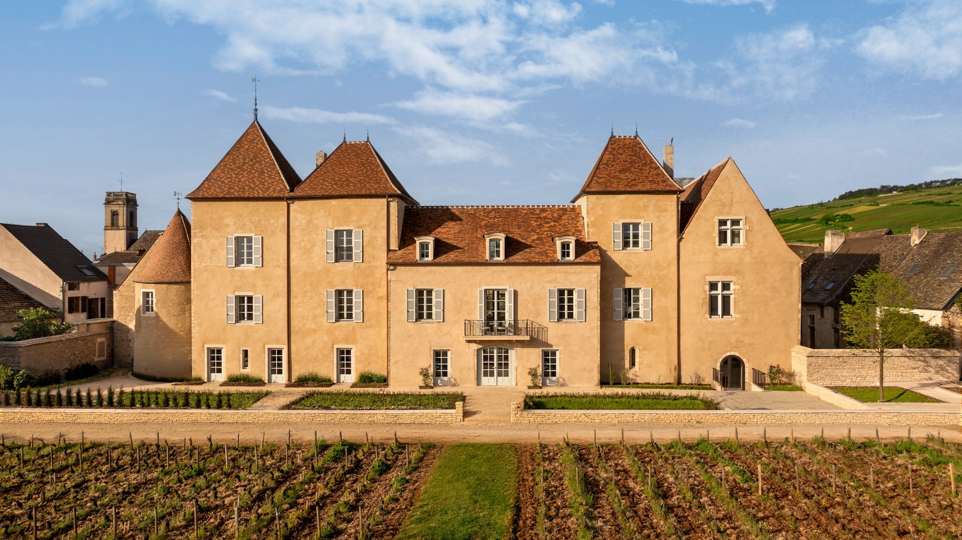 A large historic stone building with red-tiled roofs stands behind rows of vineyard vines in a rural countryside setting.