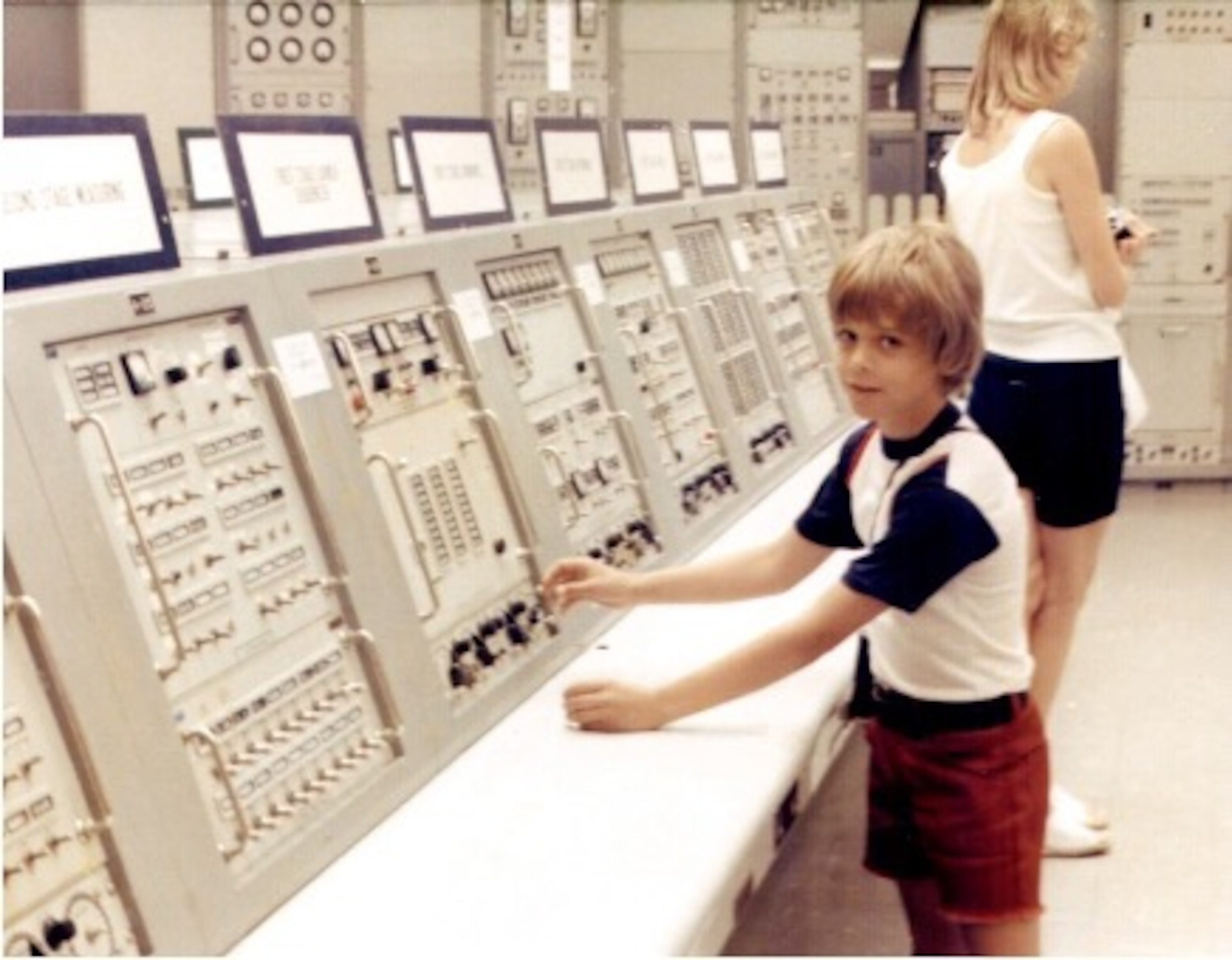 A young child in a retro control room interacts with large, complex panels filled with buttons and switches.