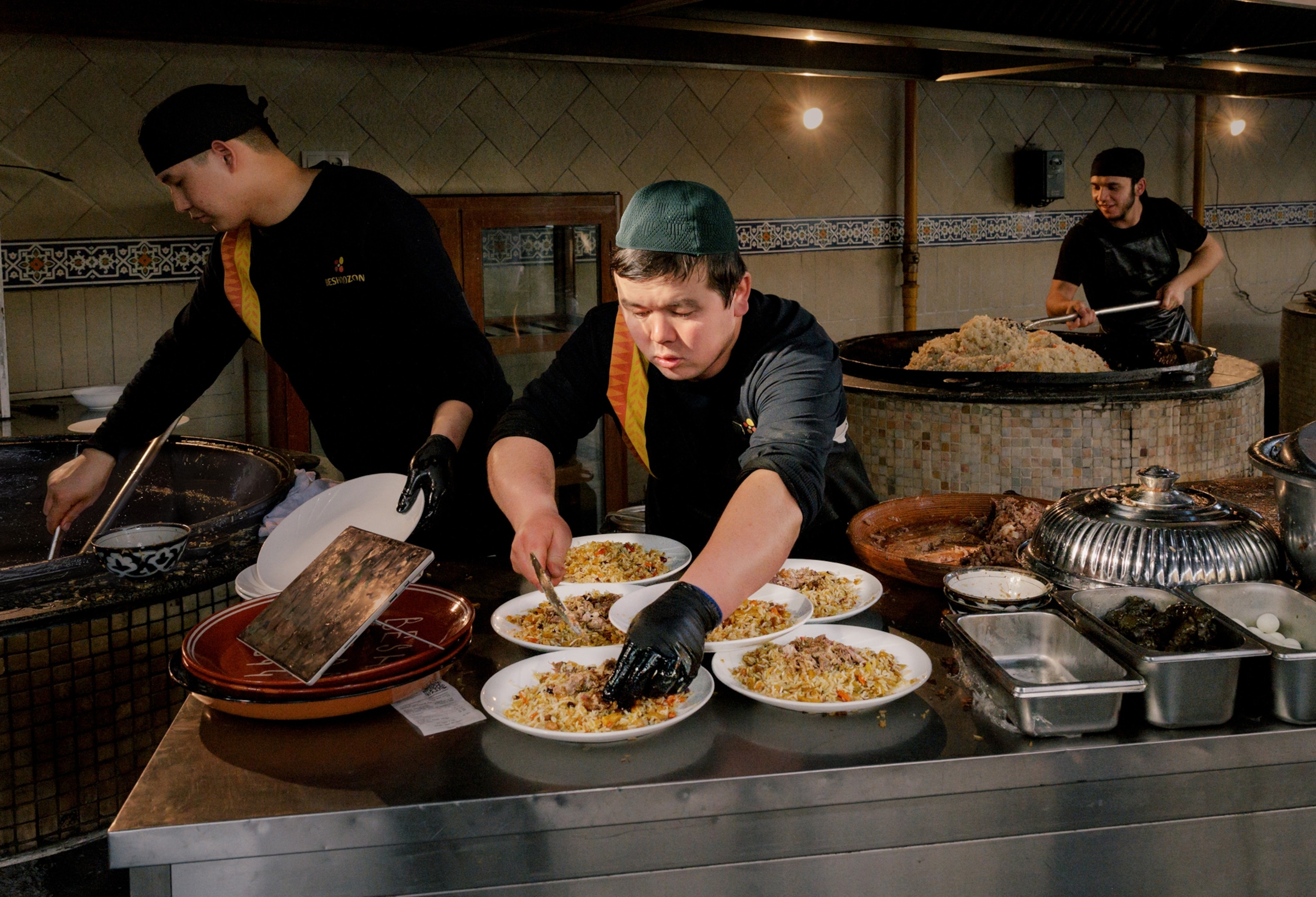 Three cooks work in a kitchen. Two people in the background make food in extremely large, round, deep cooking vessels. The man in the foreground plates food on several plates on top a metal counter.
