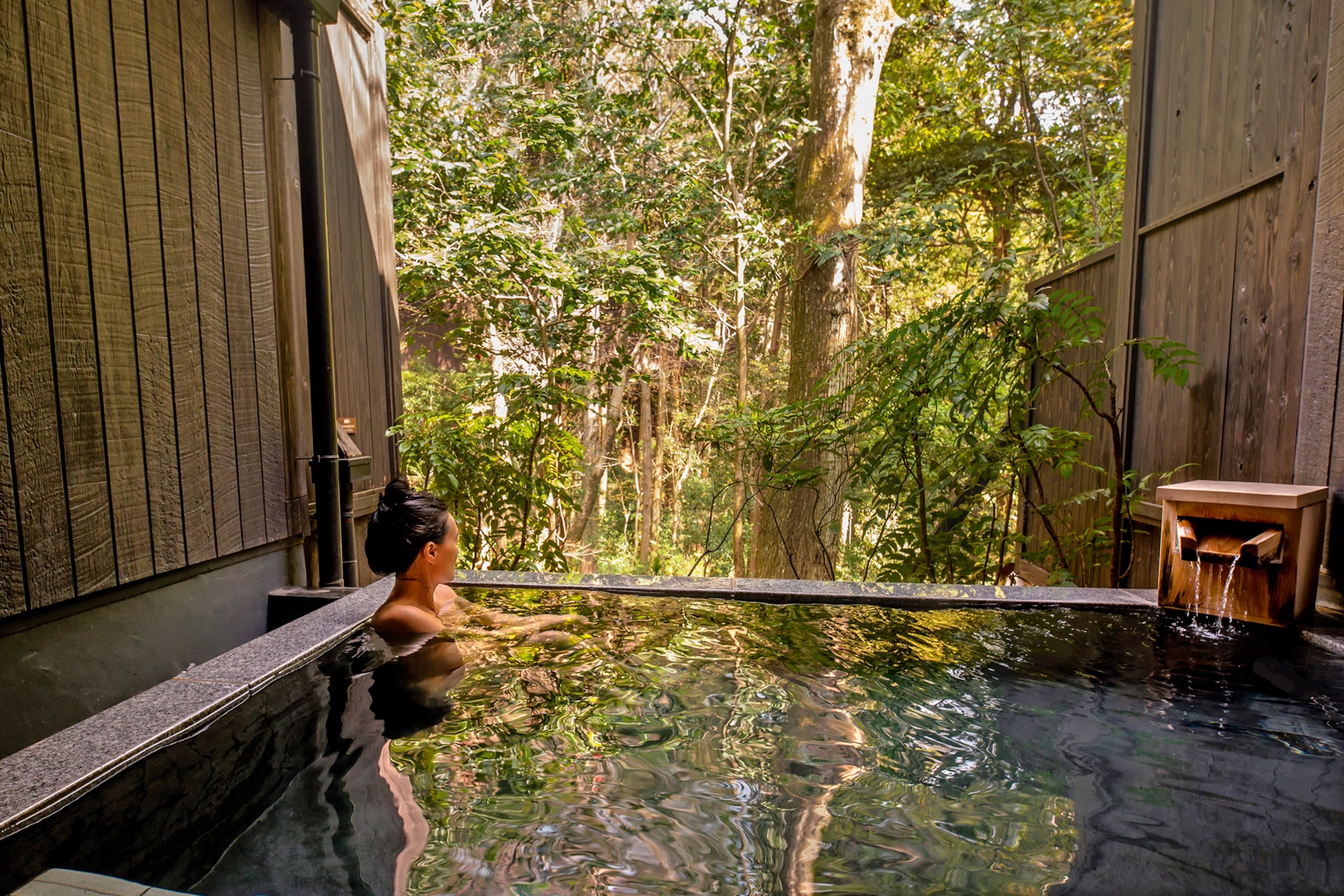 A woman with her hair up sitting in the corner of a bath looking out into the forest in front.