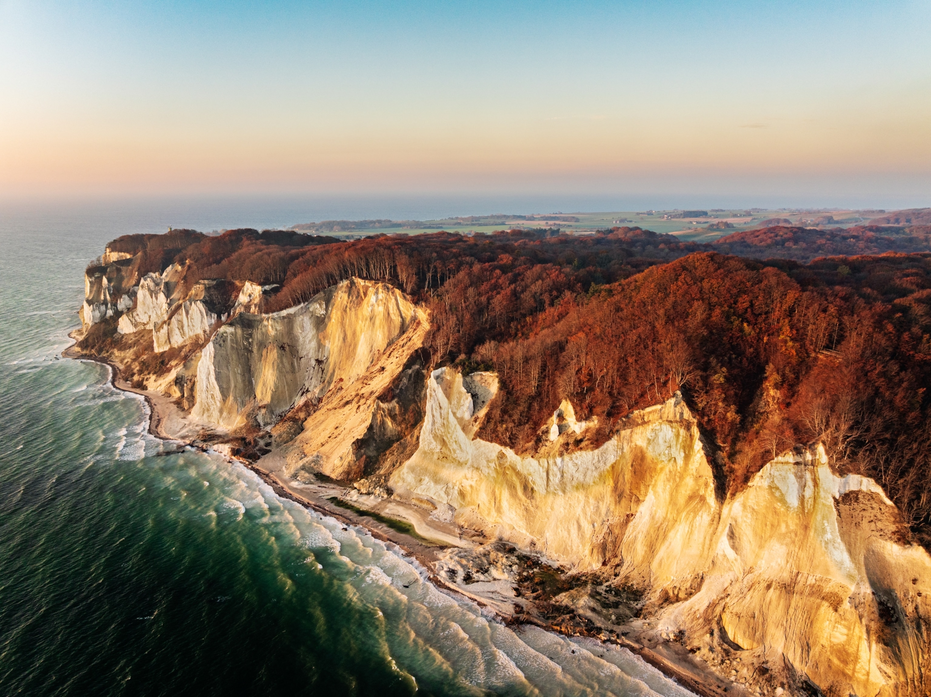 Aerial photograph of Møns Klint, Denmark, in autumn. Golden morning sunlight illuminates the white chalk cliffs, while the beech forest above displays rich orange and brown foliage. Waves break along the narrow shoreline of the Baltic Sea.