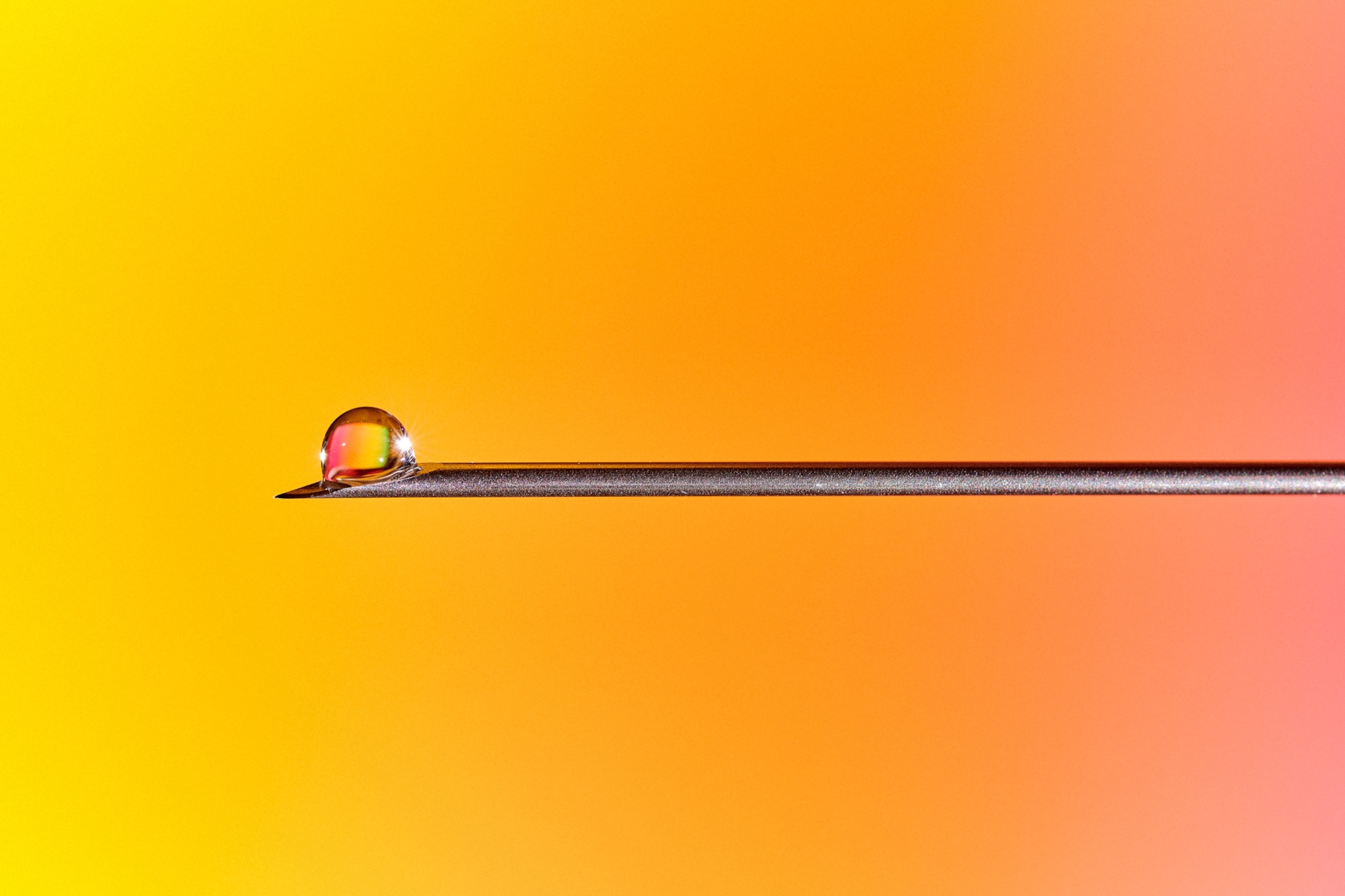 A small water droplet balances on the tip of a thin metal needle against a vivid yellow, orange, and pink gradient