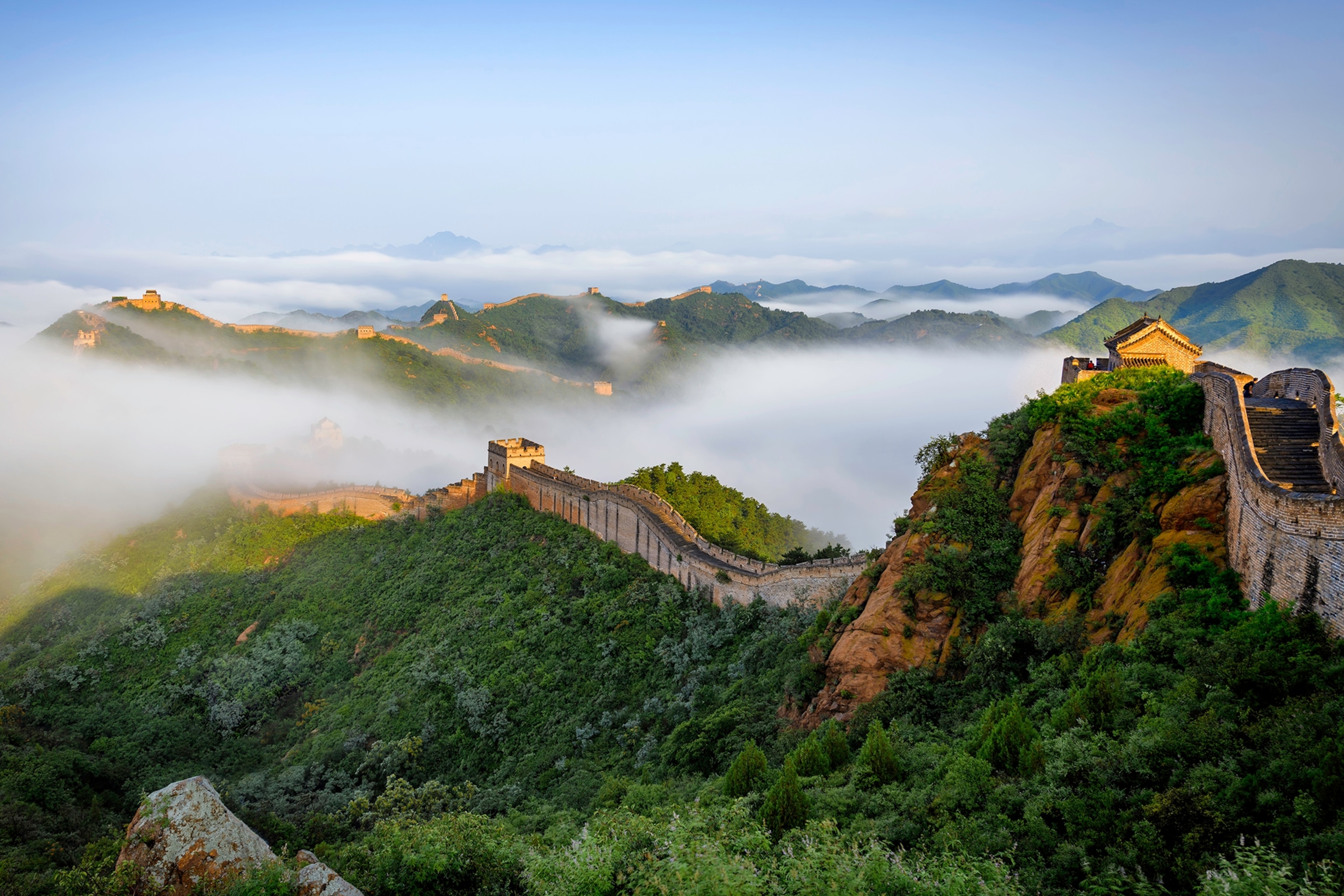 The great wall of china snaking down overgrown hills and slopes as mist spreads across mountain peaks in the distance.