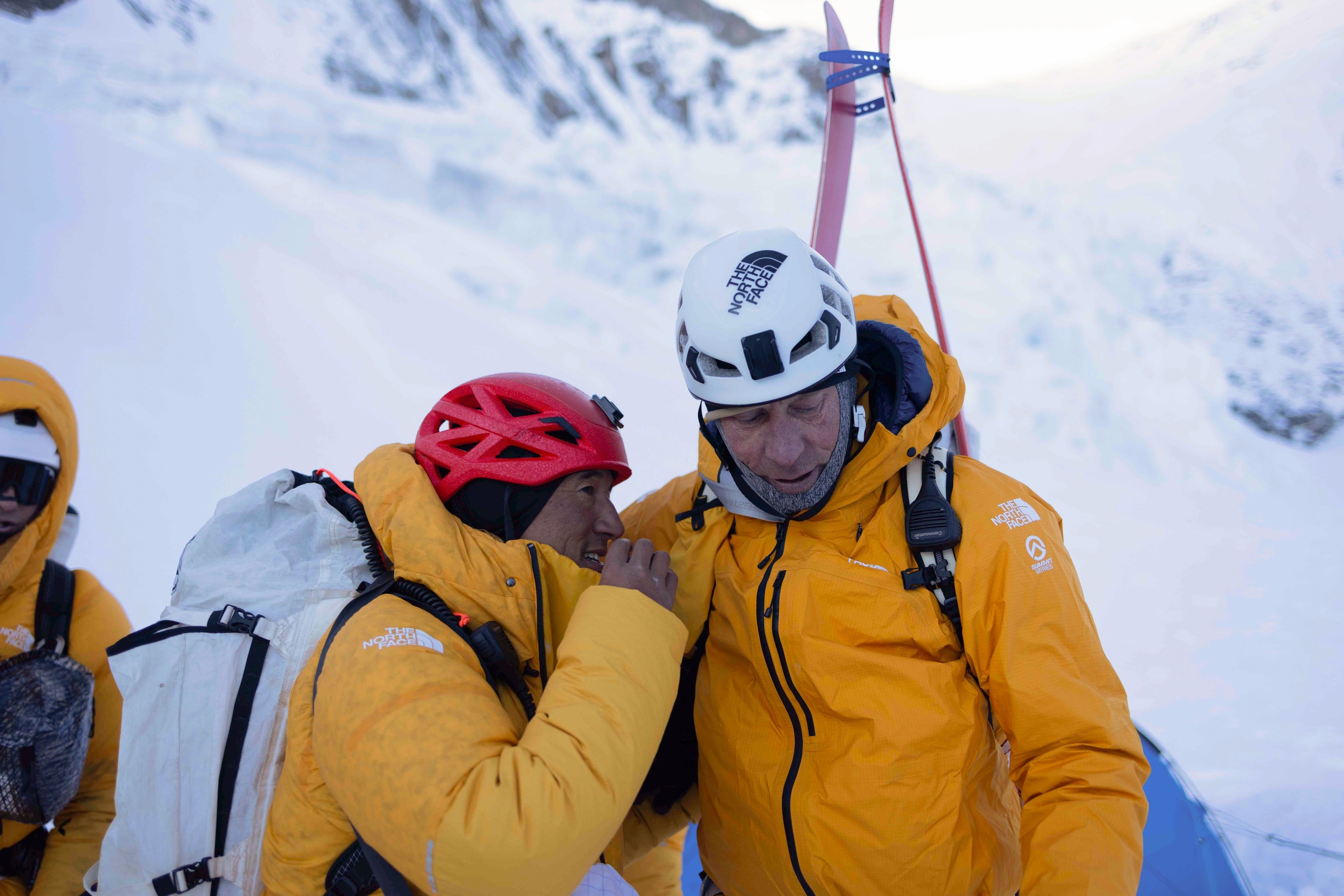 Two people wearing yellow puffer jackets talking to each other in front of a snowy background