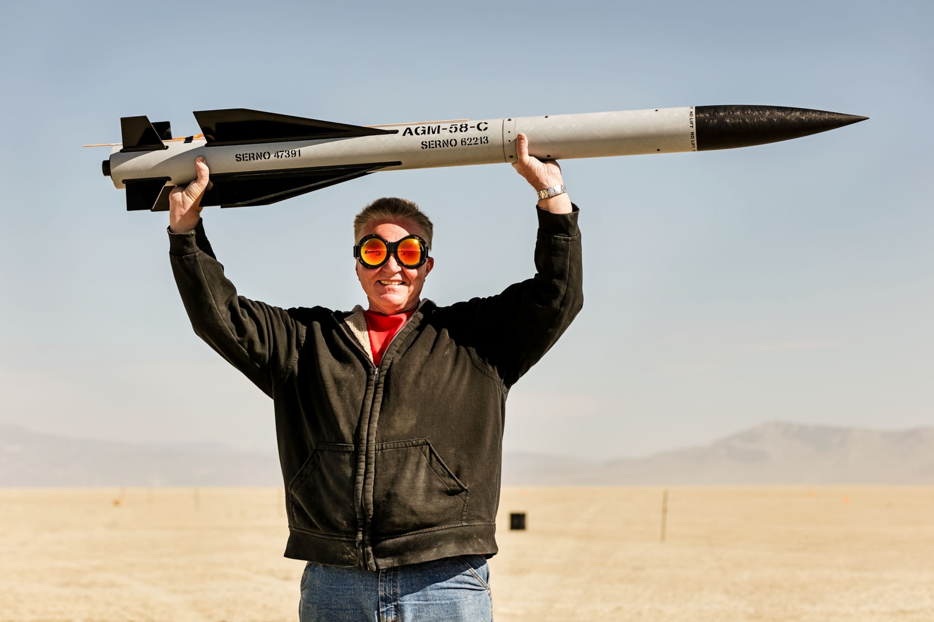 a man with orange sunglasses holding up a gray and black rocket in the desert
