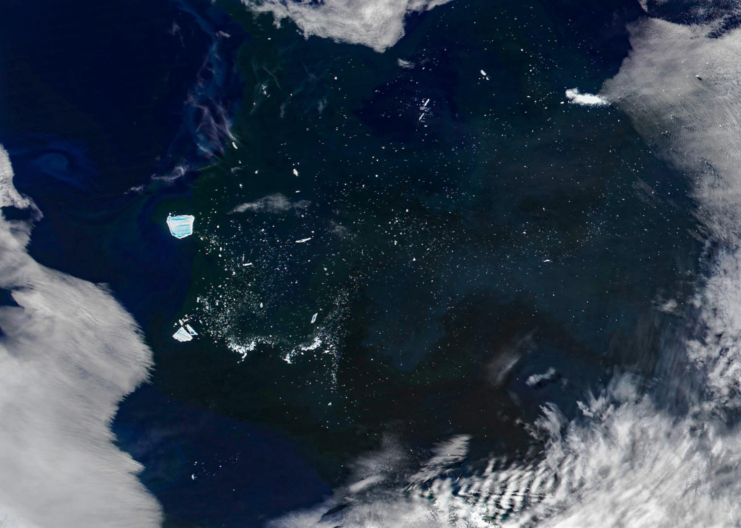 Satellite image of dark ocean with large icebergs floating near each other, surrounded by speckled smaller ice pieces. Thick cloud cover edges of the scene.