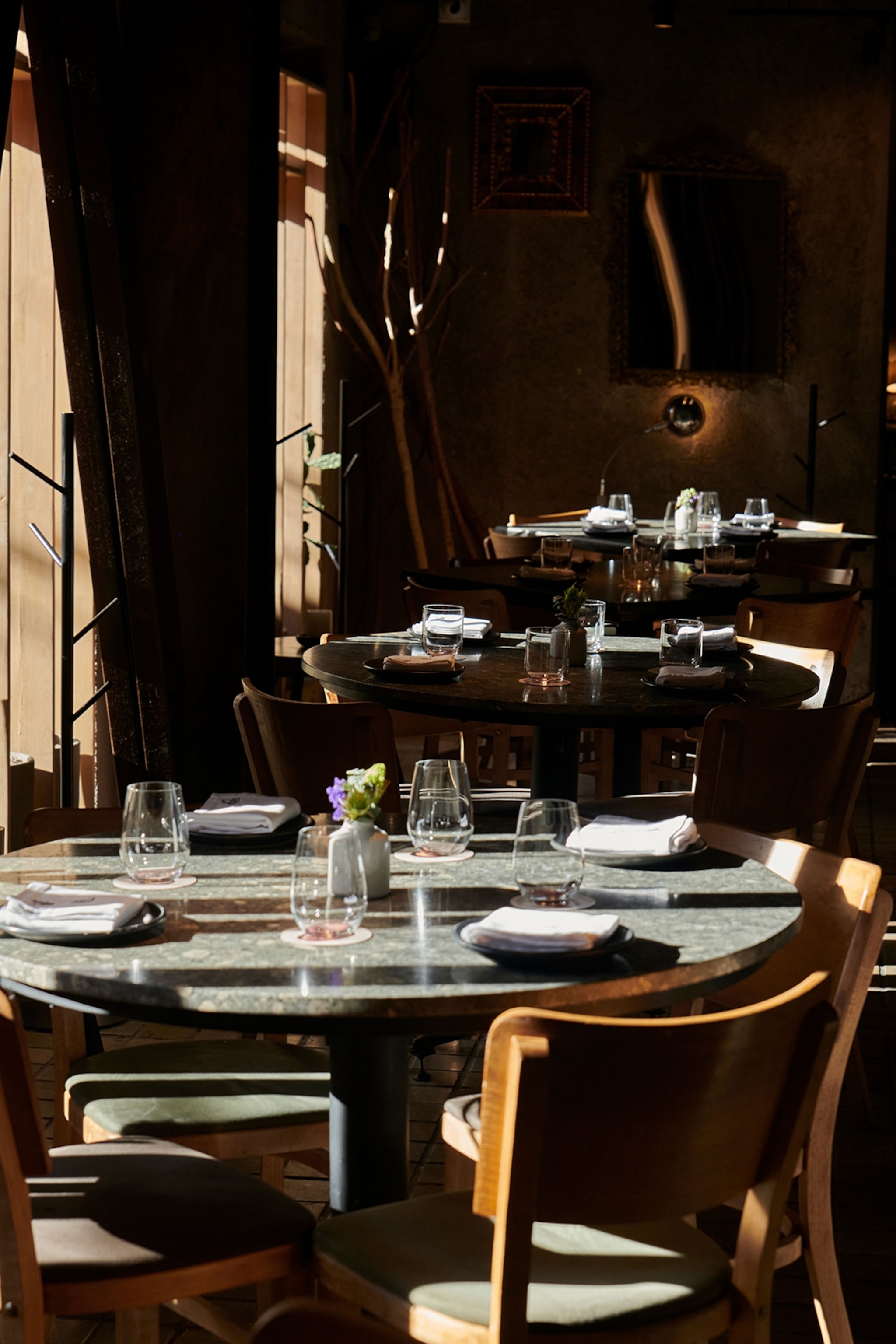 The interior of a restaurant as natural light floods through the windows and spotlights round glass tables set with linen napkins.