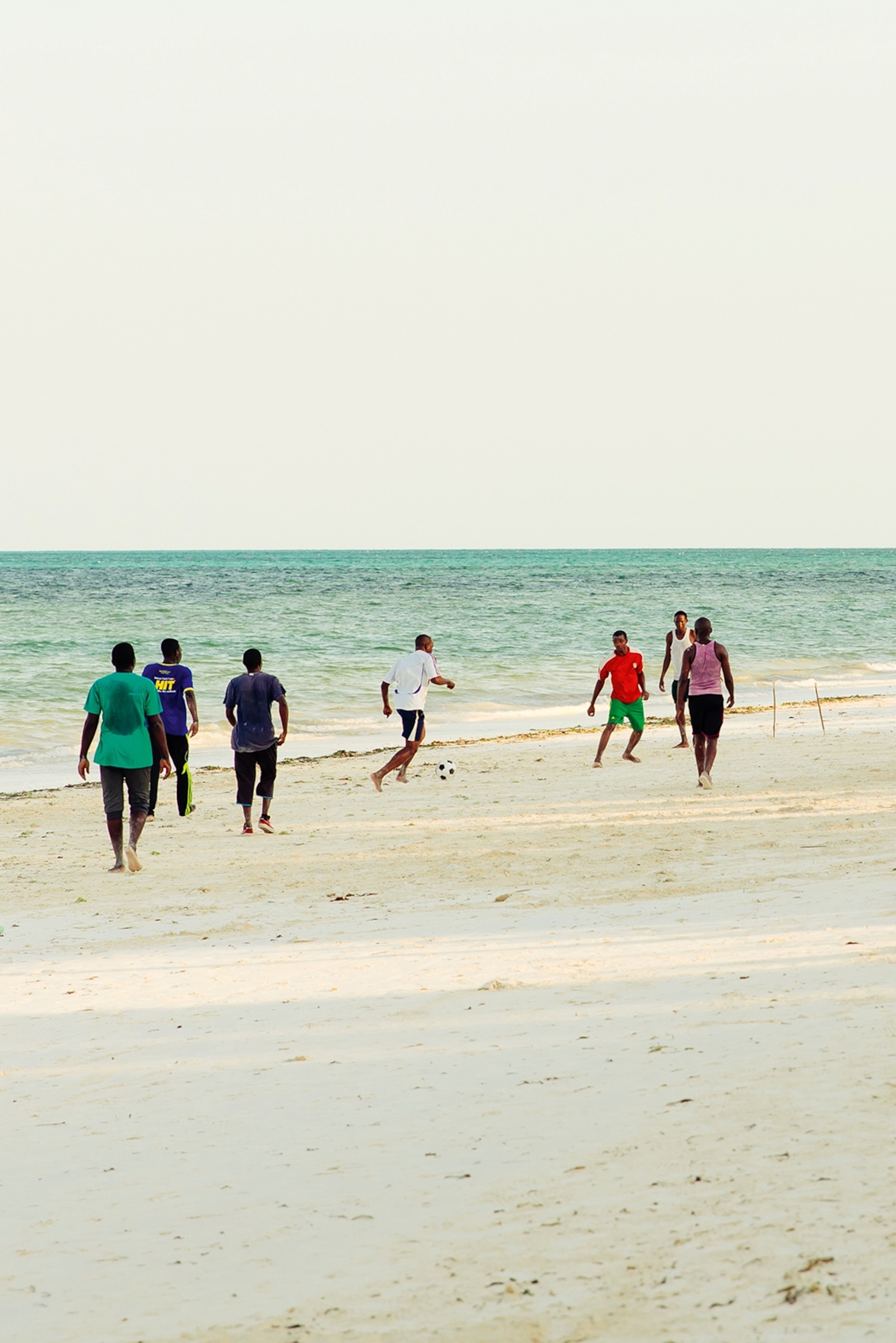 A group of young villagers playing football on a beach at early sunset.