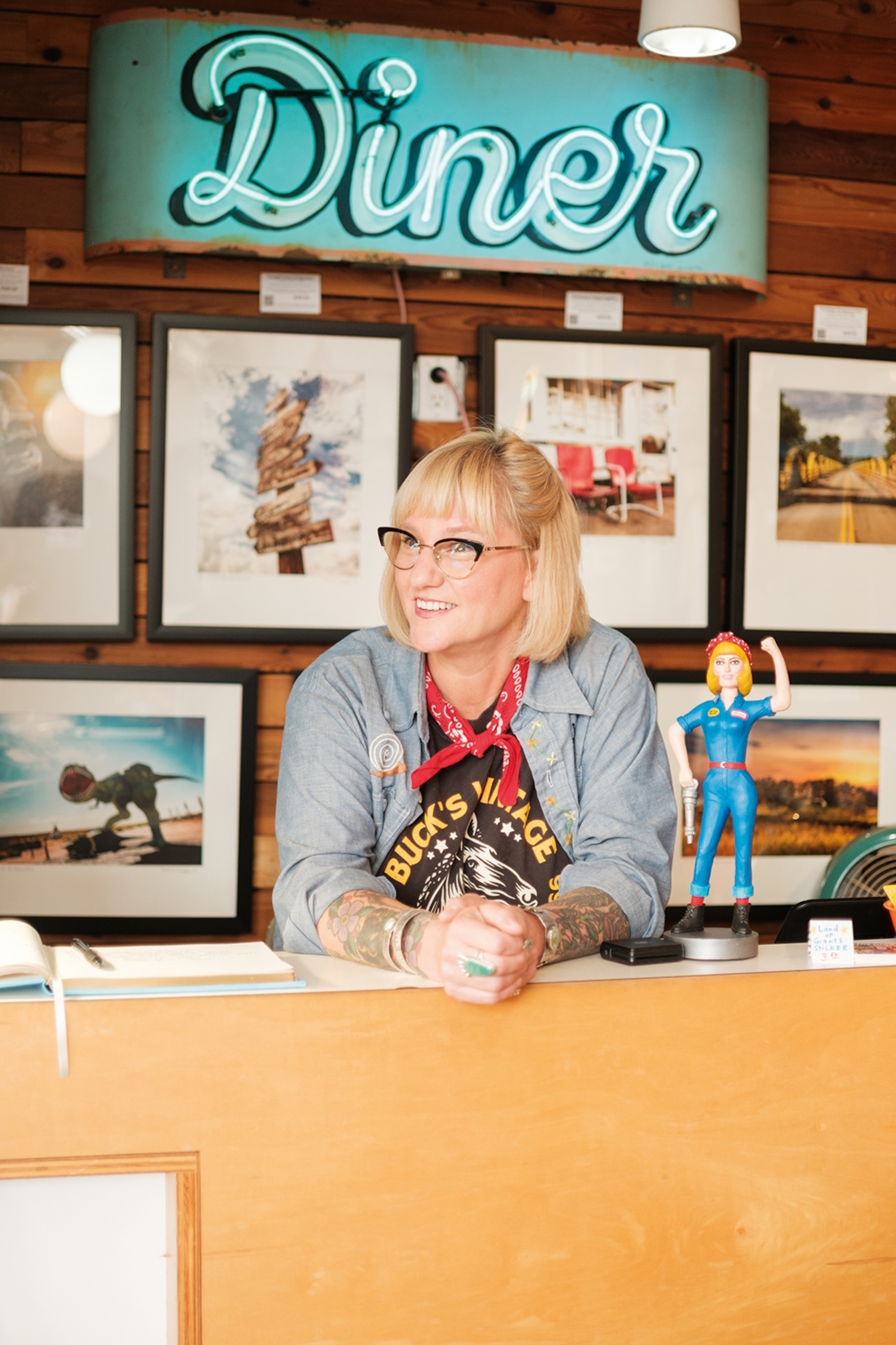 A smiling woman wearing cat-eye-rimmed glasses and a red handkerchief around her neck, leaning onto the counter of her shop.