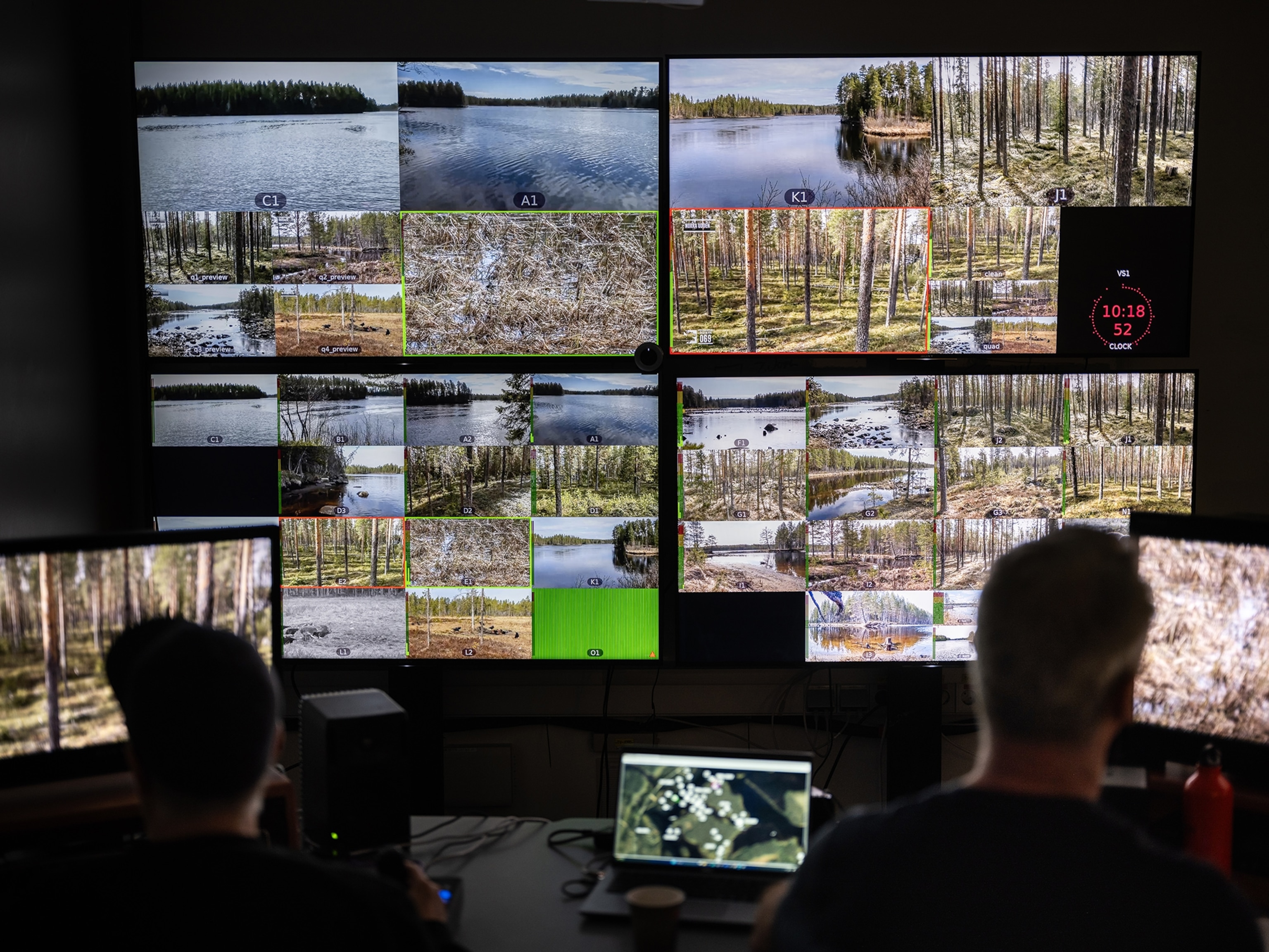 two men are seen from behind in front of a wall of TV screens showing nature scenes