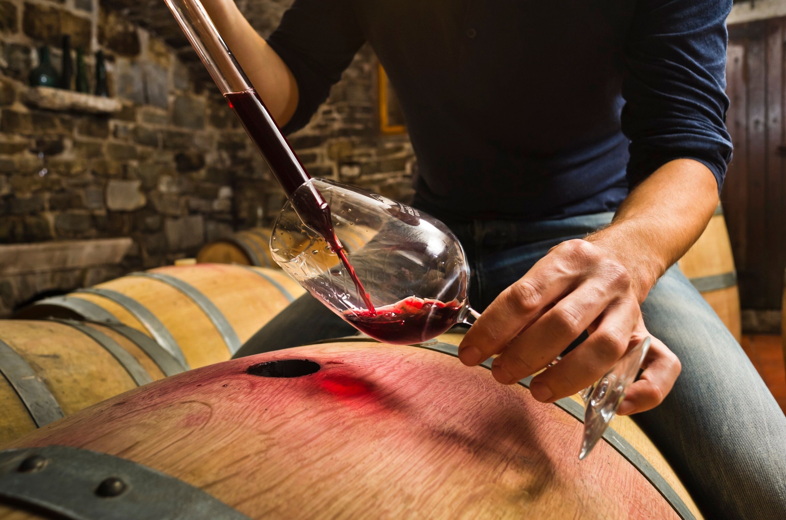 A person pours a glass of red wine. Light shines through it, casting a wooden cask in a red hue..