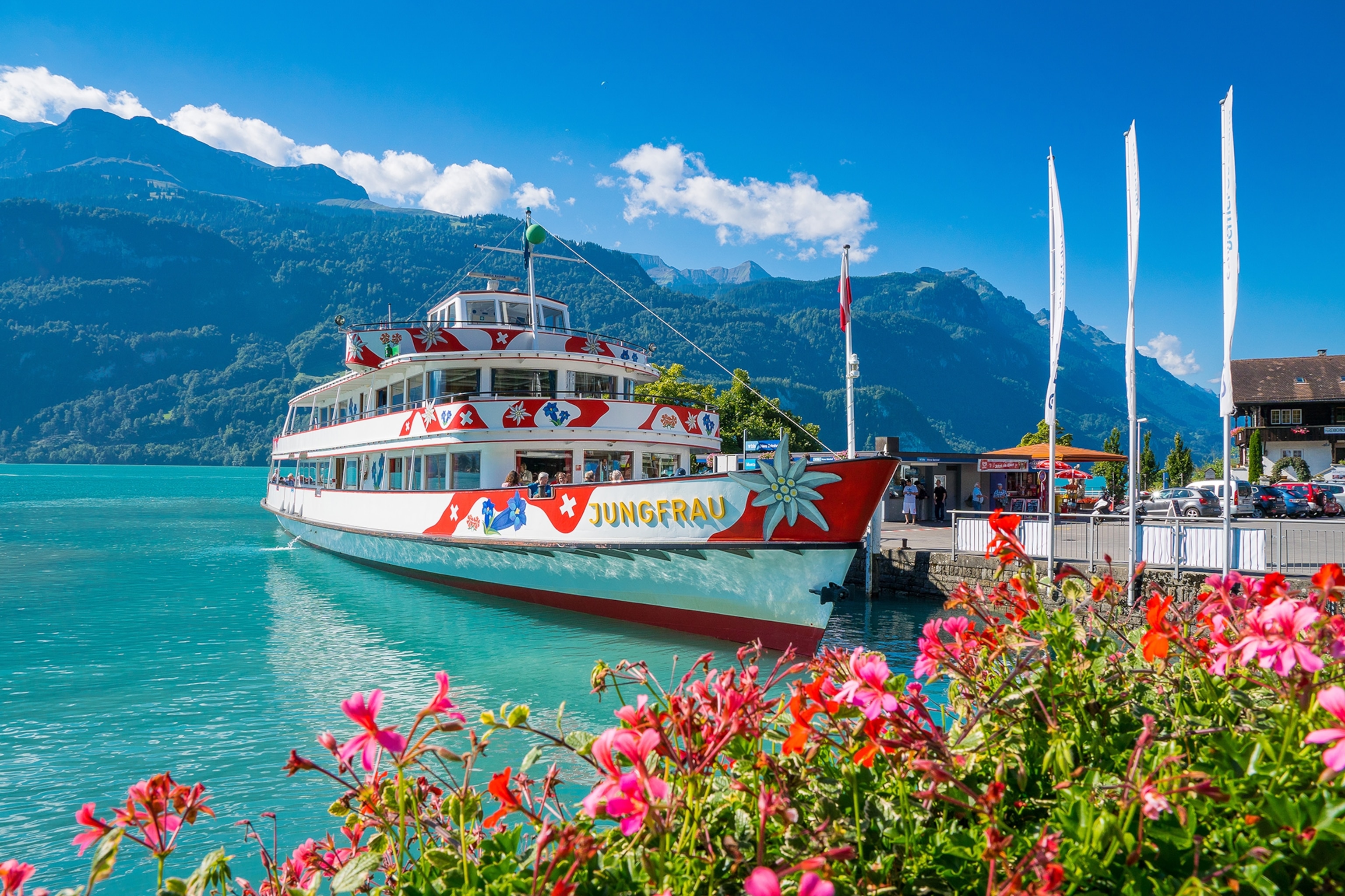 A white boat with red and blue decorations sits at a dock on the shore of Lake Brienz.