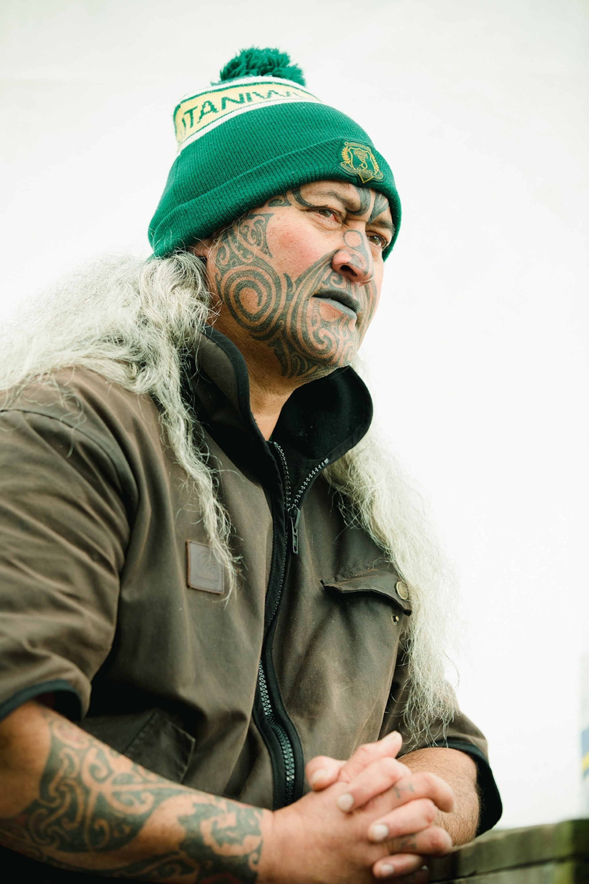 A portrait of a male Māori elder with traditional face tattoos and long, wavy, white hair.