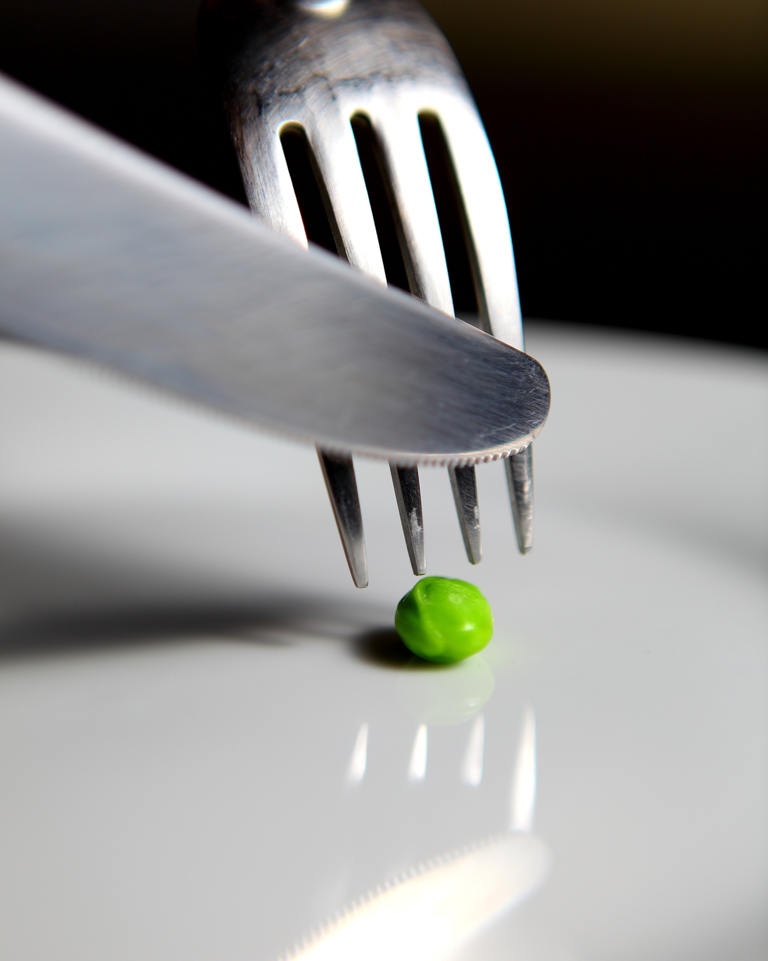 A single green pea on a white plate is placed between a fork and a knife