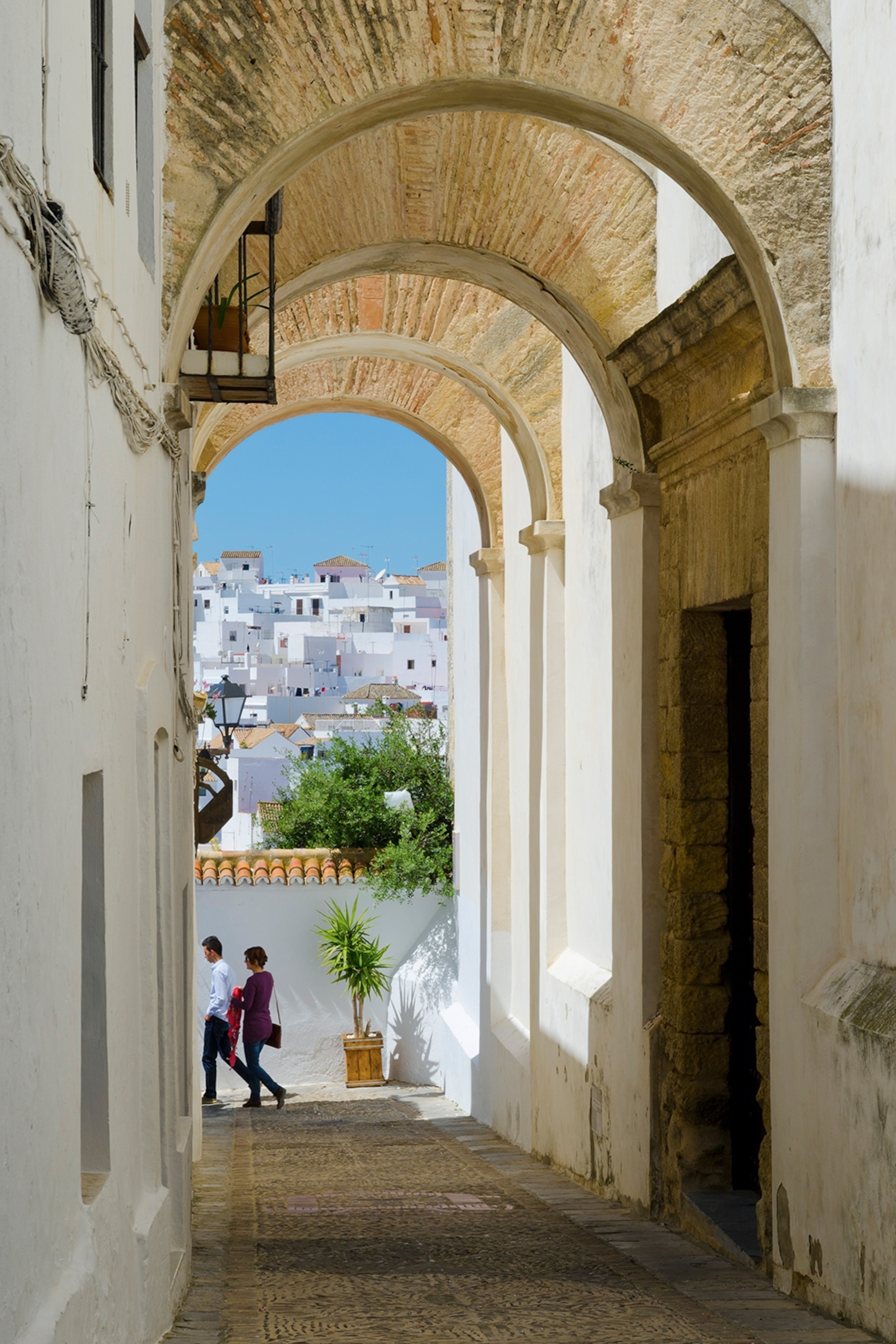 A narrow and arched alleyway leading into an opening overlooking a hill-side town.