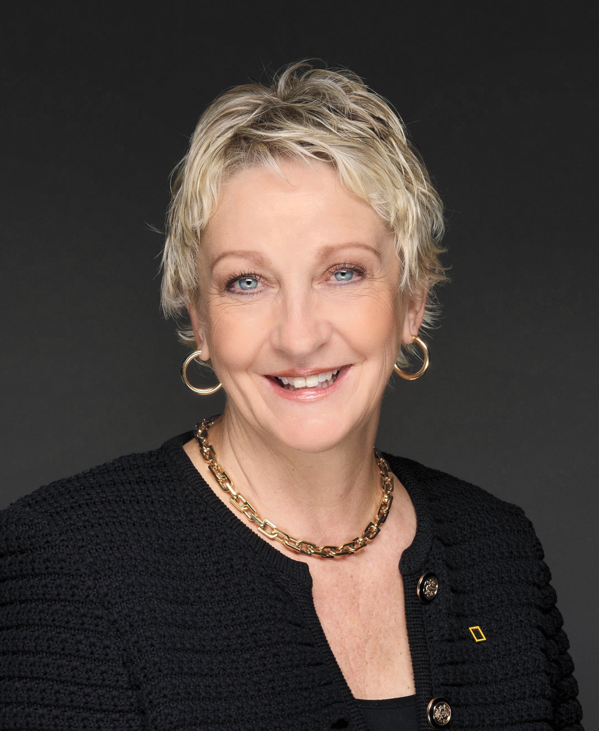 Portrait of a smiling woman with short blondish heir in black dress with golden neckless and loop earrings.
