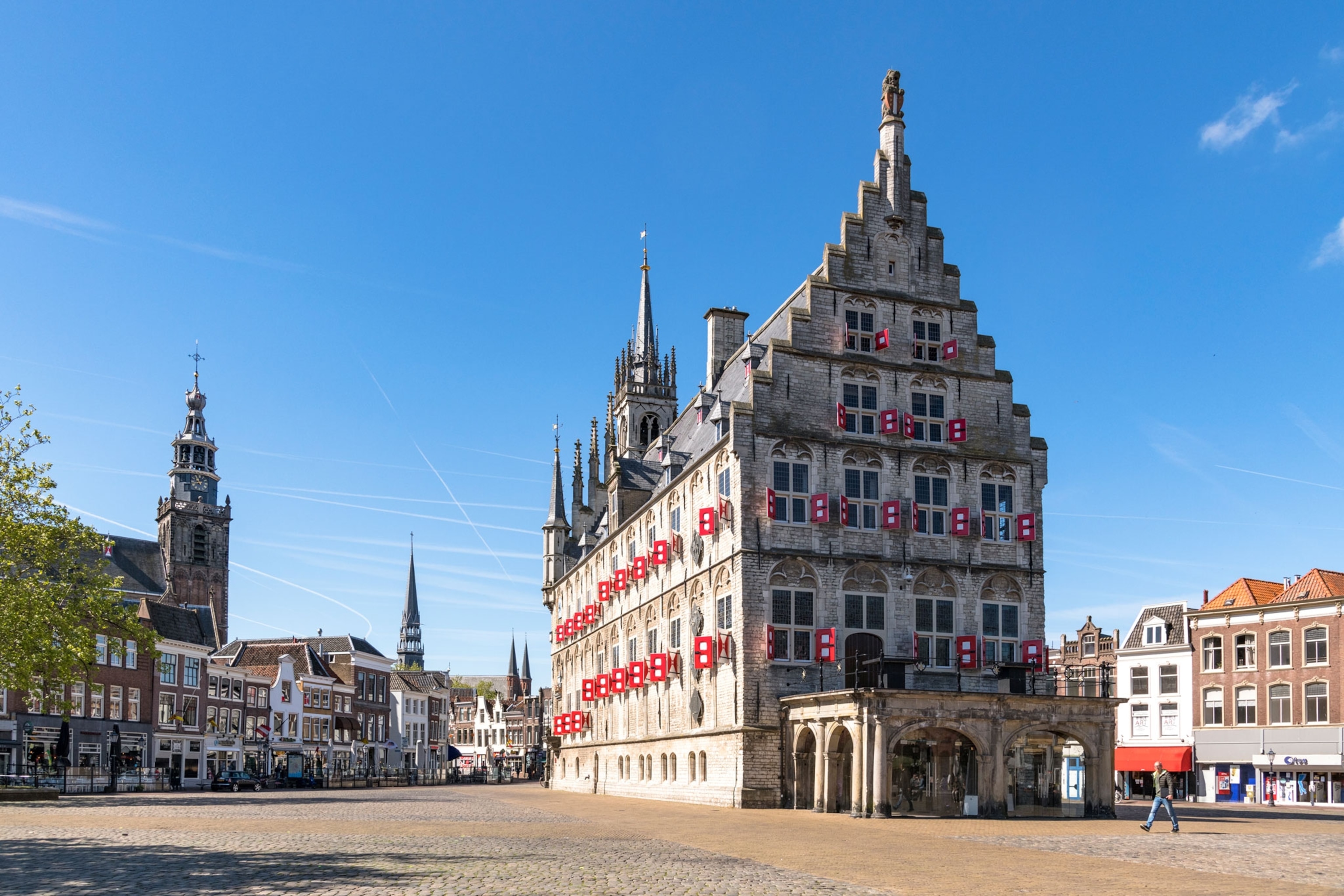 A historic stone building with stepped gables and red-and-white shutters stands on a cobblestone square under a clear blue sky, surrounded by smaller buildings and church towers in the background.