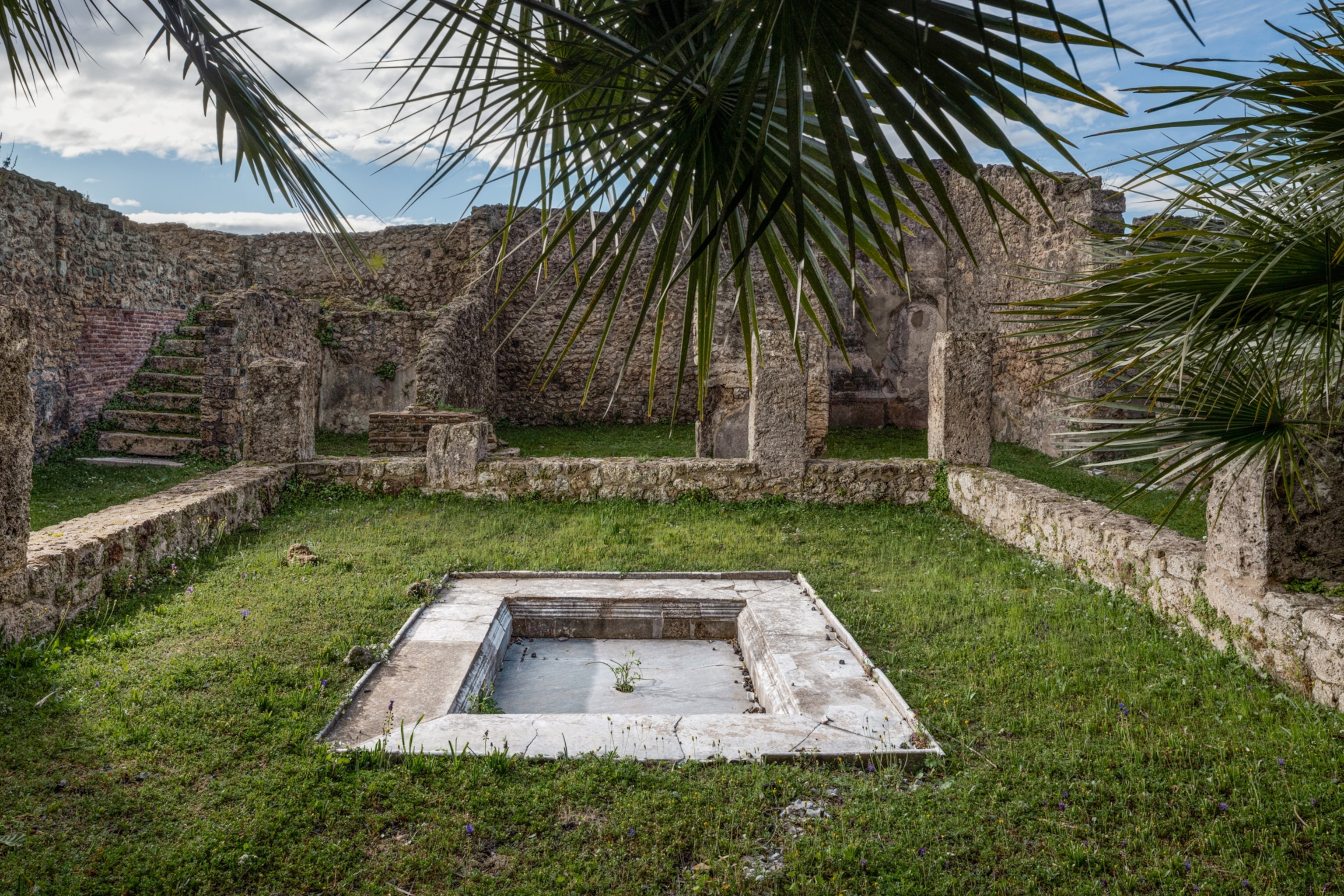 Ancient stone ruins in Pompeii featuring a central rectangular marble basin surrounded by grass, stone walls, and palm fronds against a blue sky.