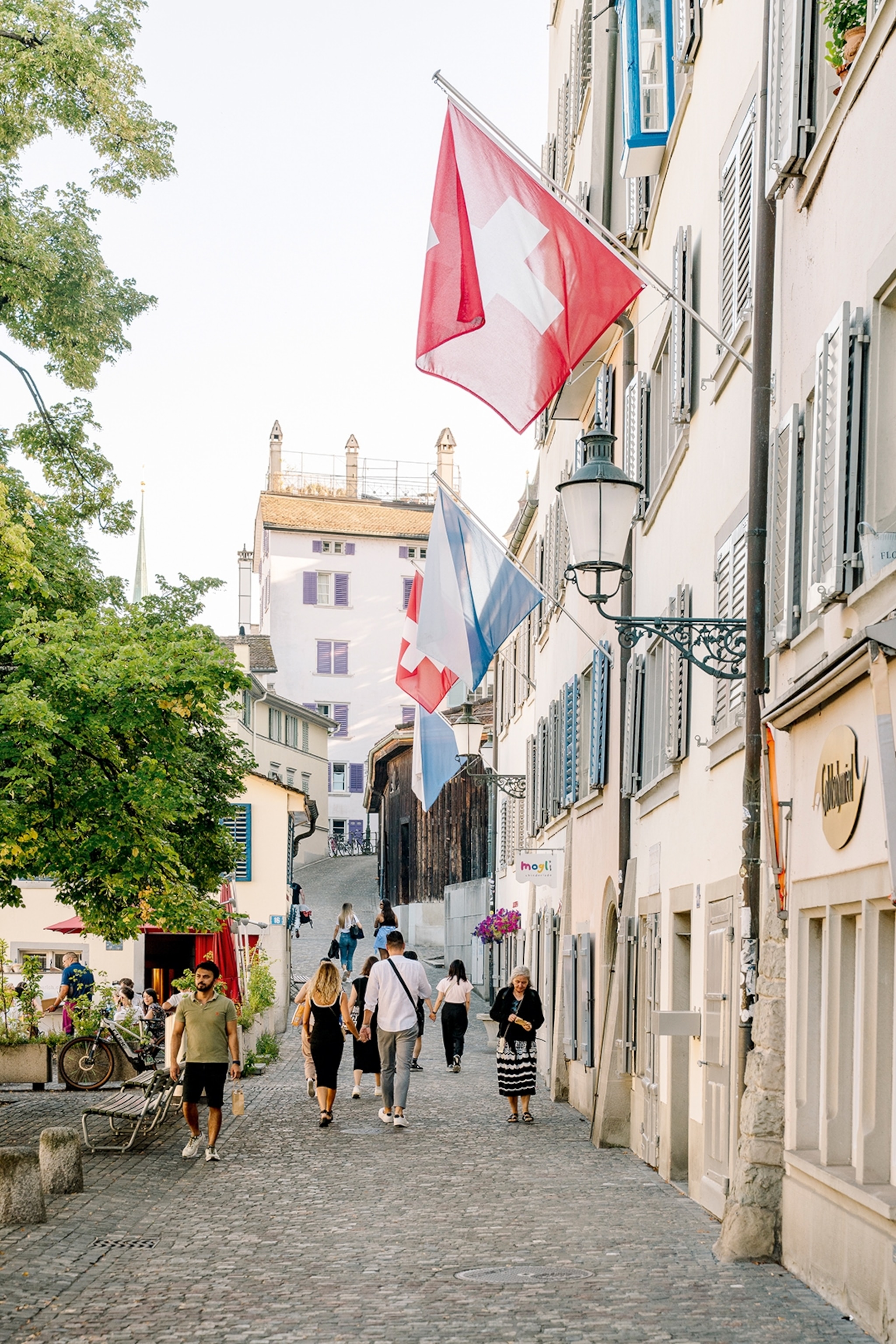 A calm, cobble-stoned alleyway with Swiss flags hung along one building's wall and a small courtyard with trees opposite.