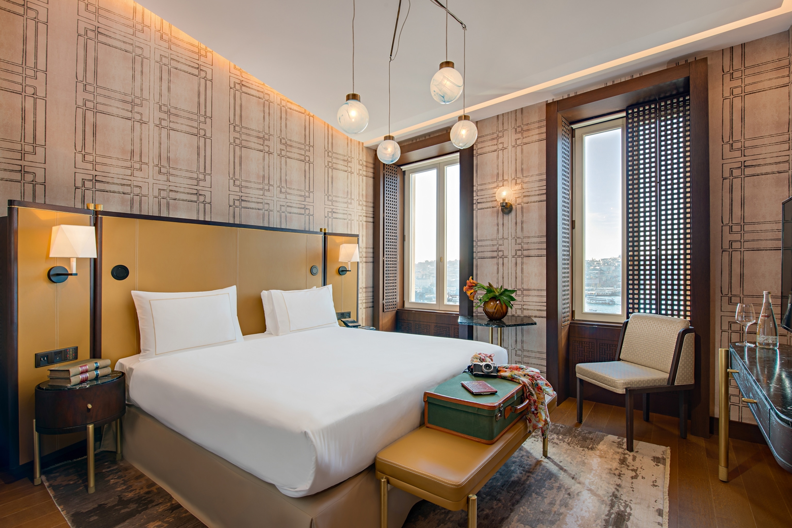 A cosy queen-sized hotel bedroom with a tasteful, graphic wallpaper, simple globe chandelier and a tall window showcasing the edge of the hill-top city.