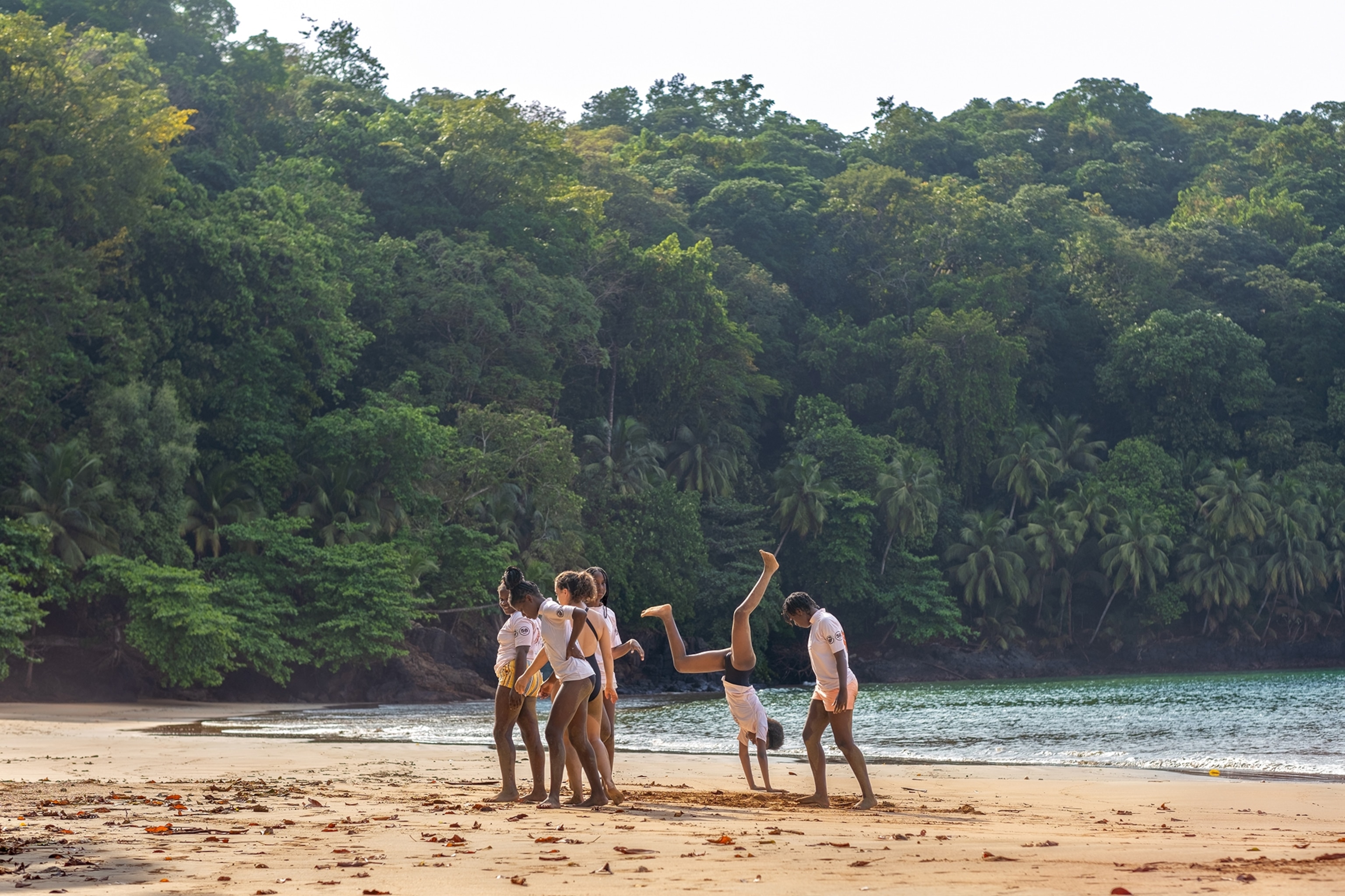 A group of young kids playing on the beach, doing cartwheels in the sand, with a rainforest in the background.
