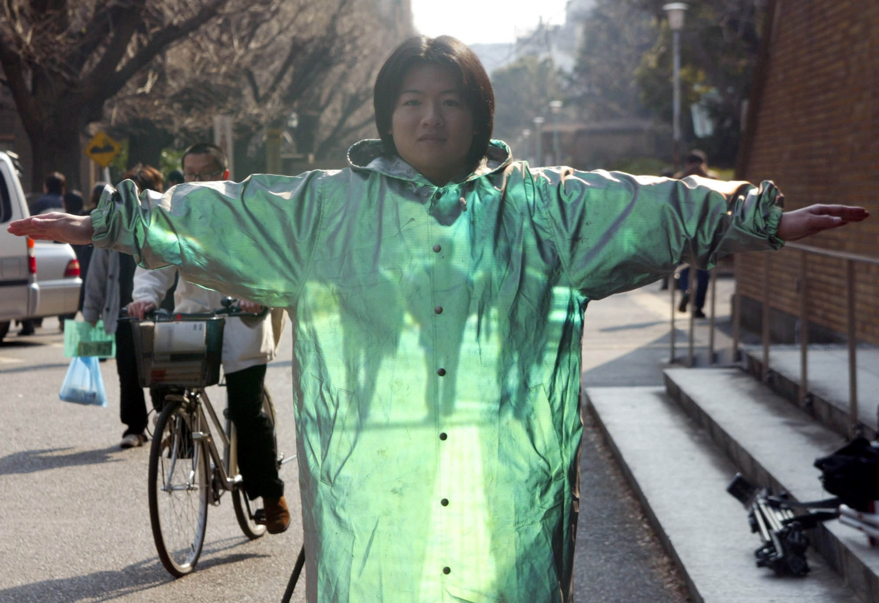 A person stands with arms outstretched, wearing a shiny, translucent green coat that appears to make the wearing partially invisible.