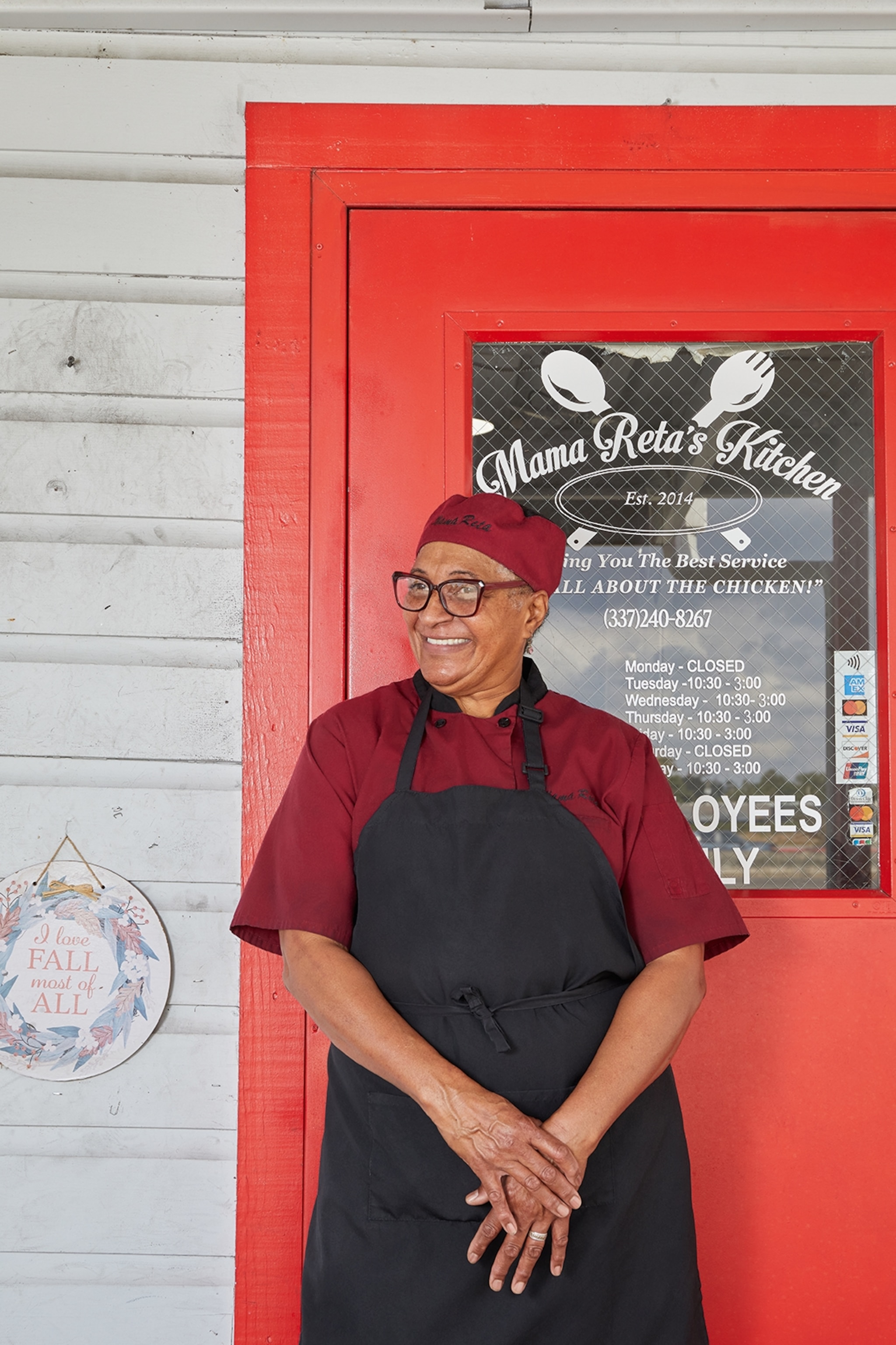 A smiling elderly woman wearing an apron in front of her BBQ restaurant.