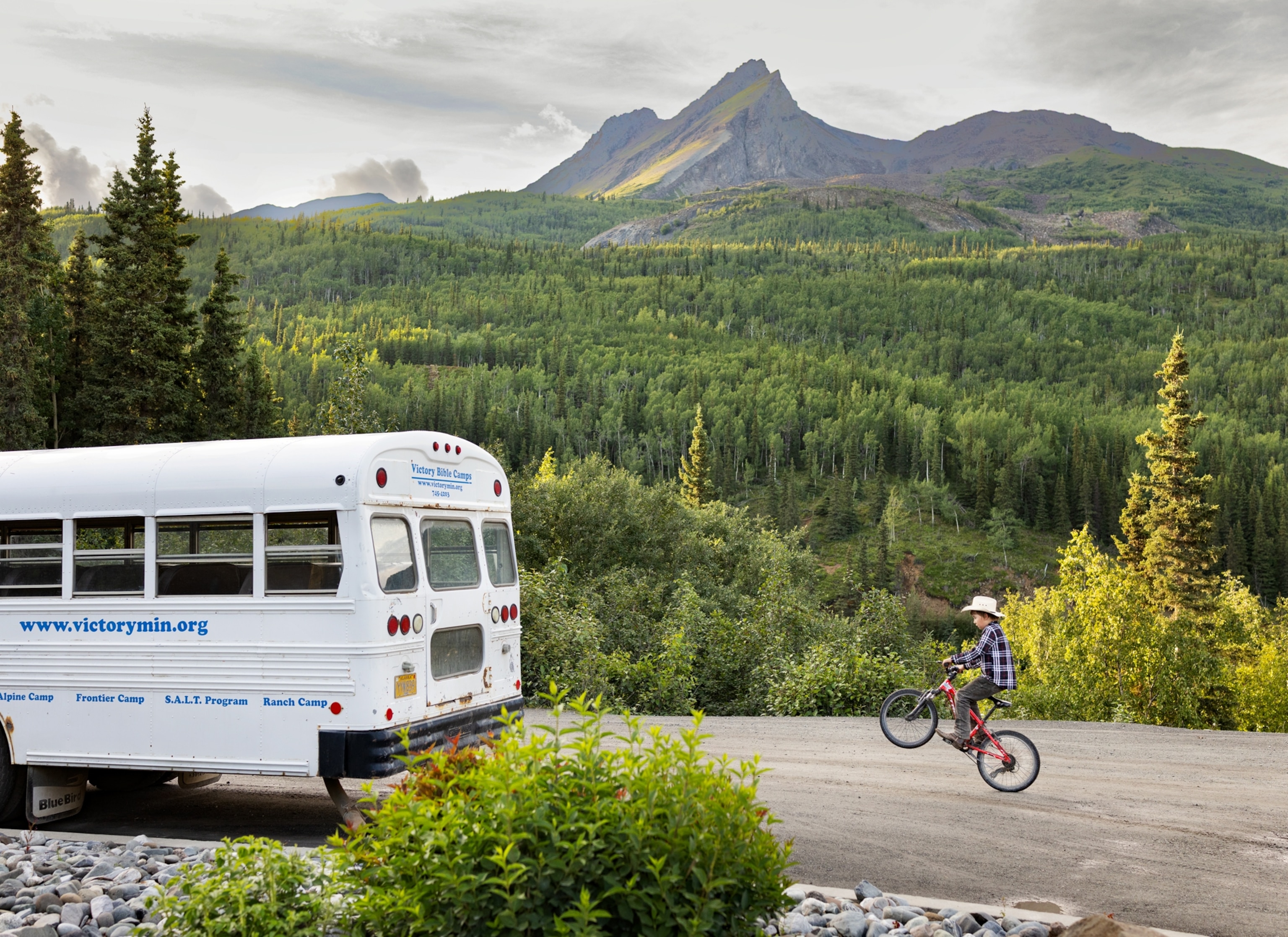 A white bus is parked on a dirt road while a cyclist rides past it, with green forest and tall mountains in the background.